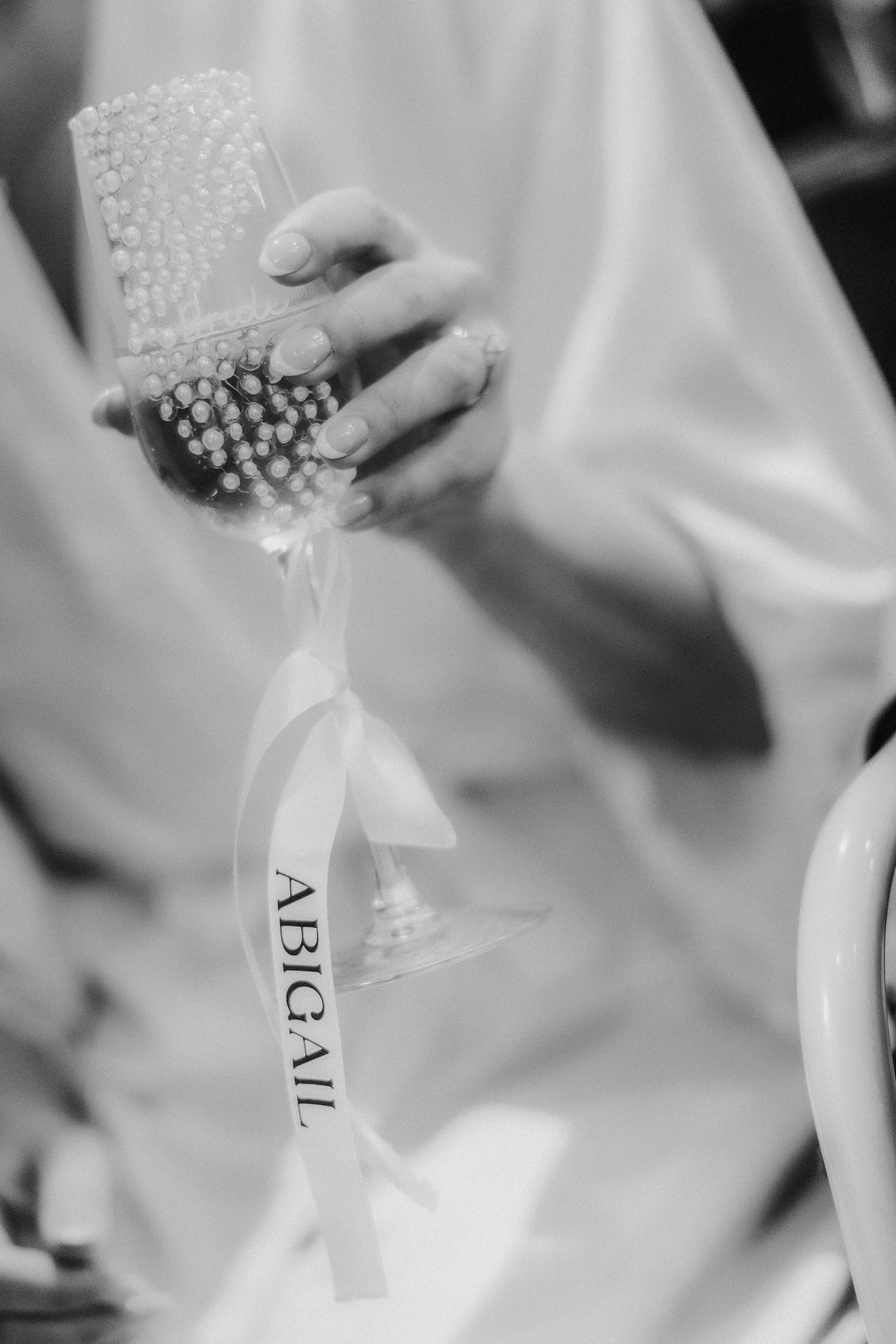 Hand holding a pearl-studded champagne flute with a ribbon reading 'ABIGAIL'.