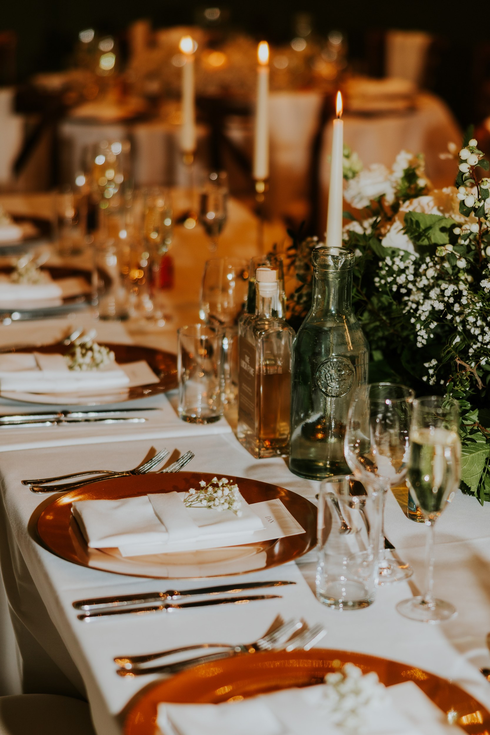 Laid wedding reception table with charger plates, napkins, glassware, bottles, lit taper candles and a floral centerpiece.