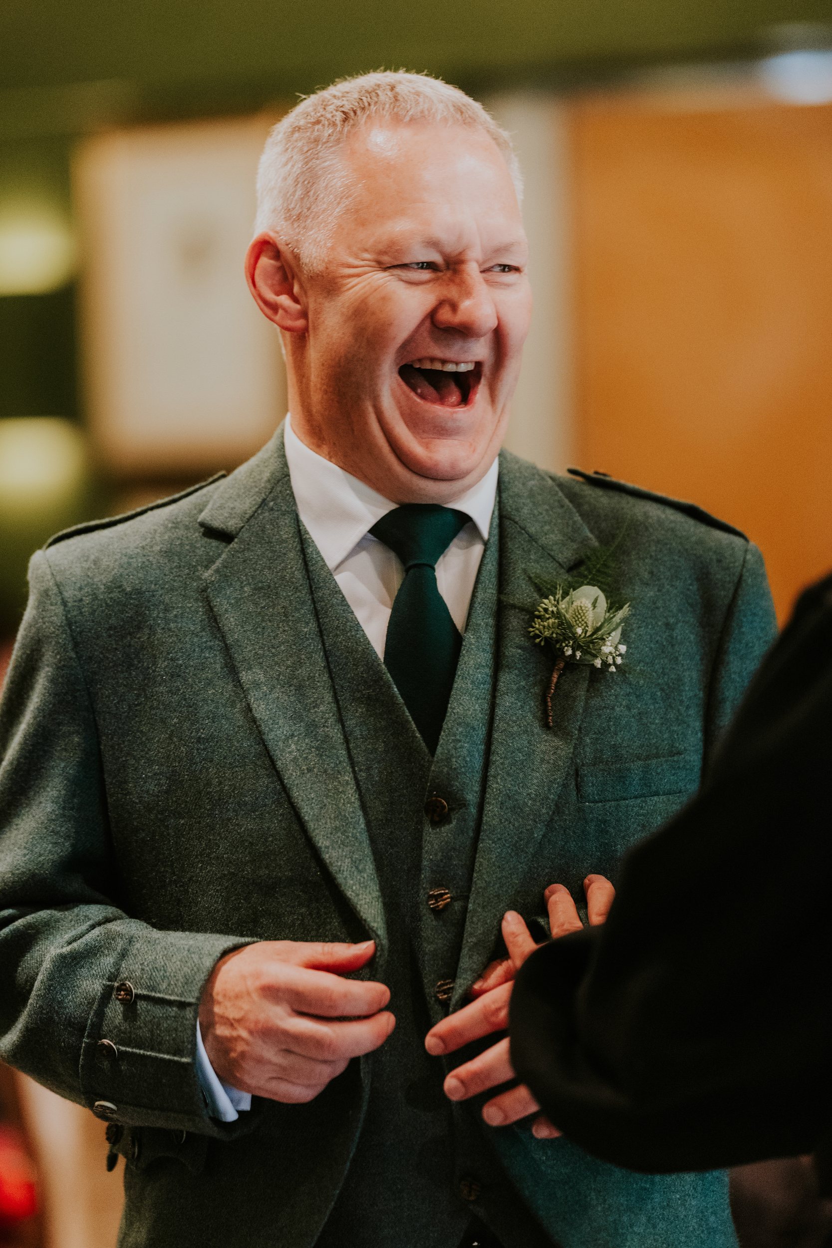 Laughing man in green tweed suit with boutonniere, indoor close-up showing hands.