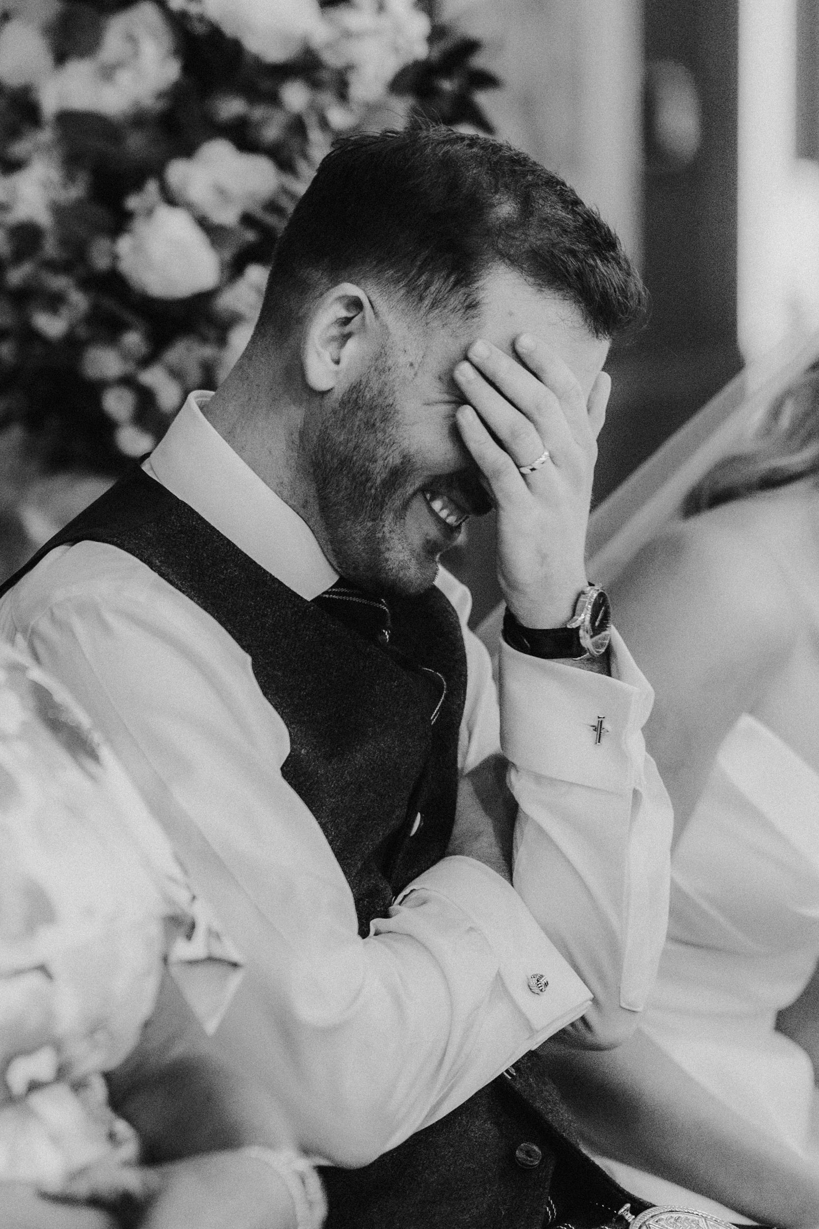 Man in waistcoat laughing with his hand over his face at a wedding reception table.