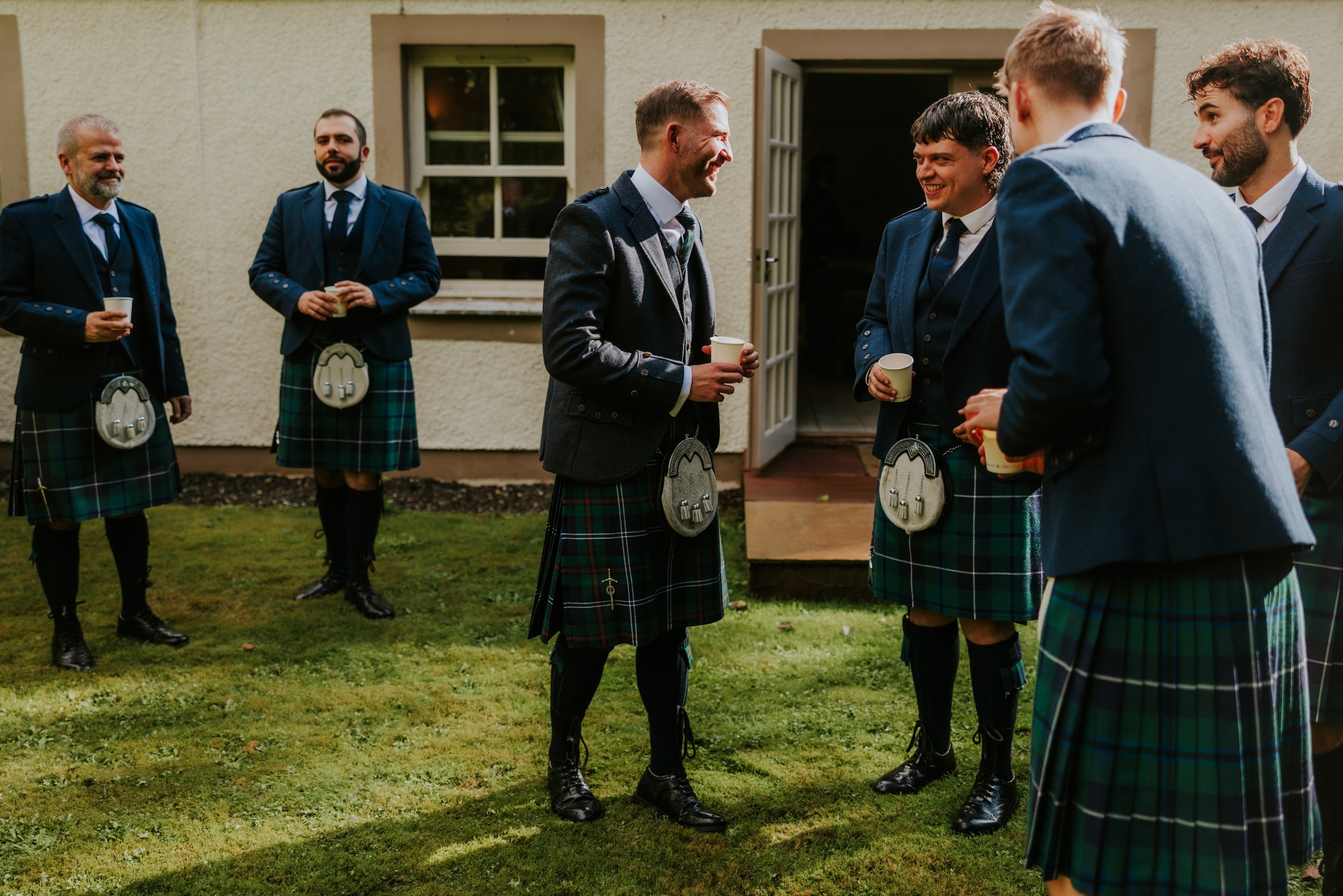 Men in kilts chatting and holding paper cups outside a pale building on the lawn.