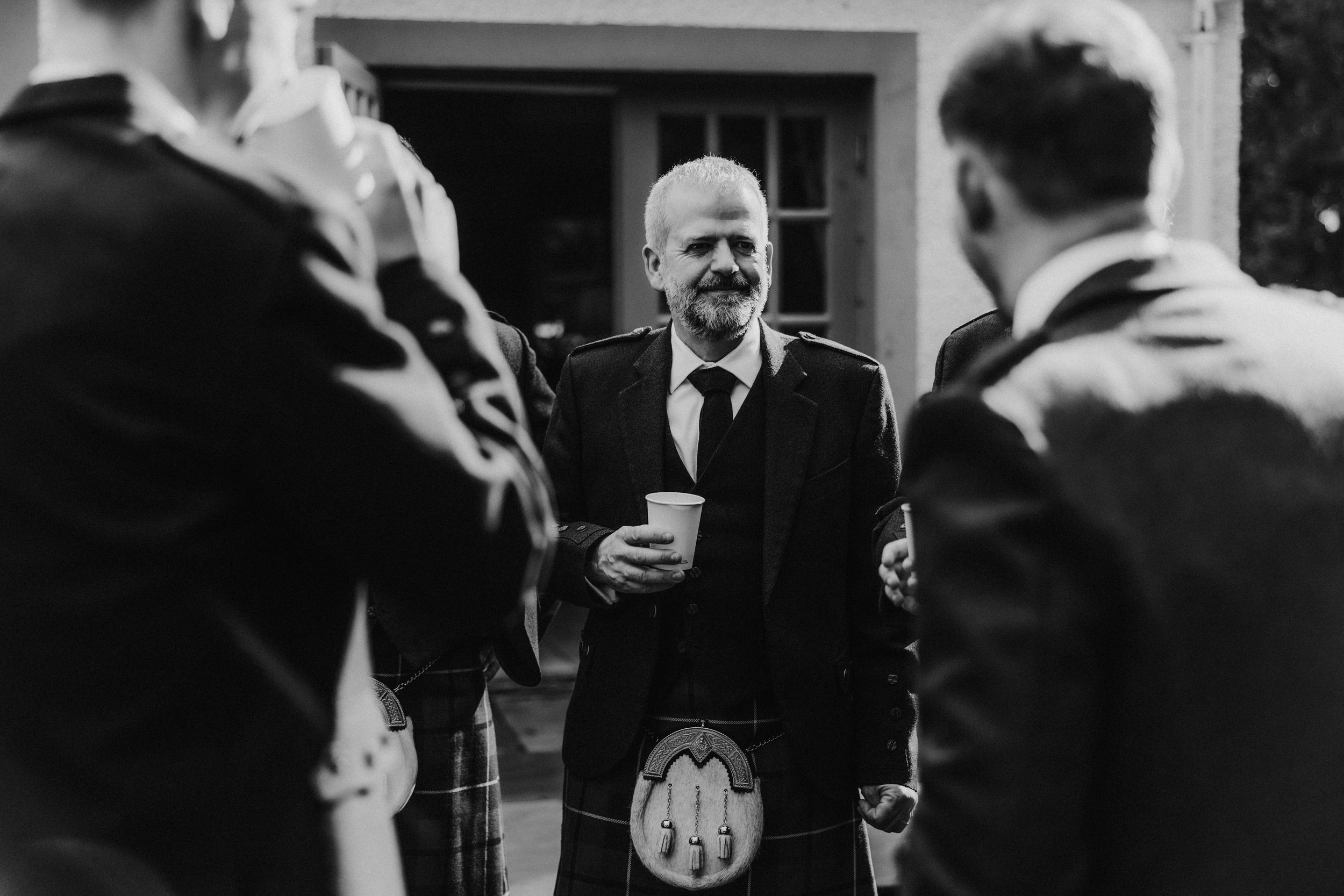 Men in kilts chatting outside, centre bearded man holding a paper cup.