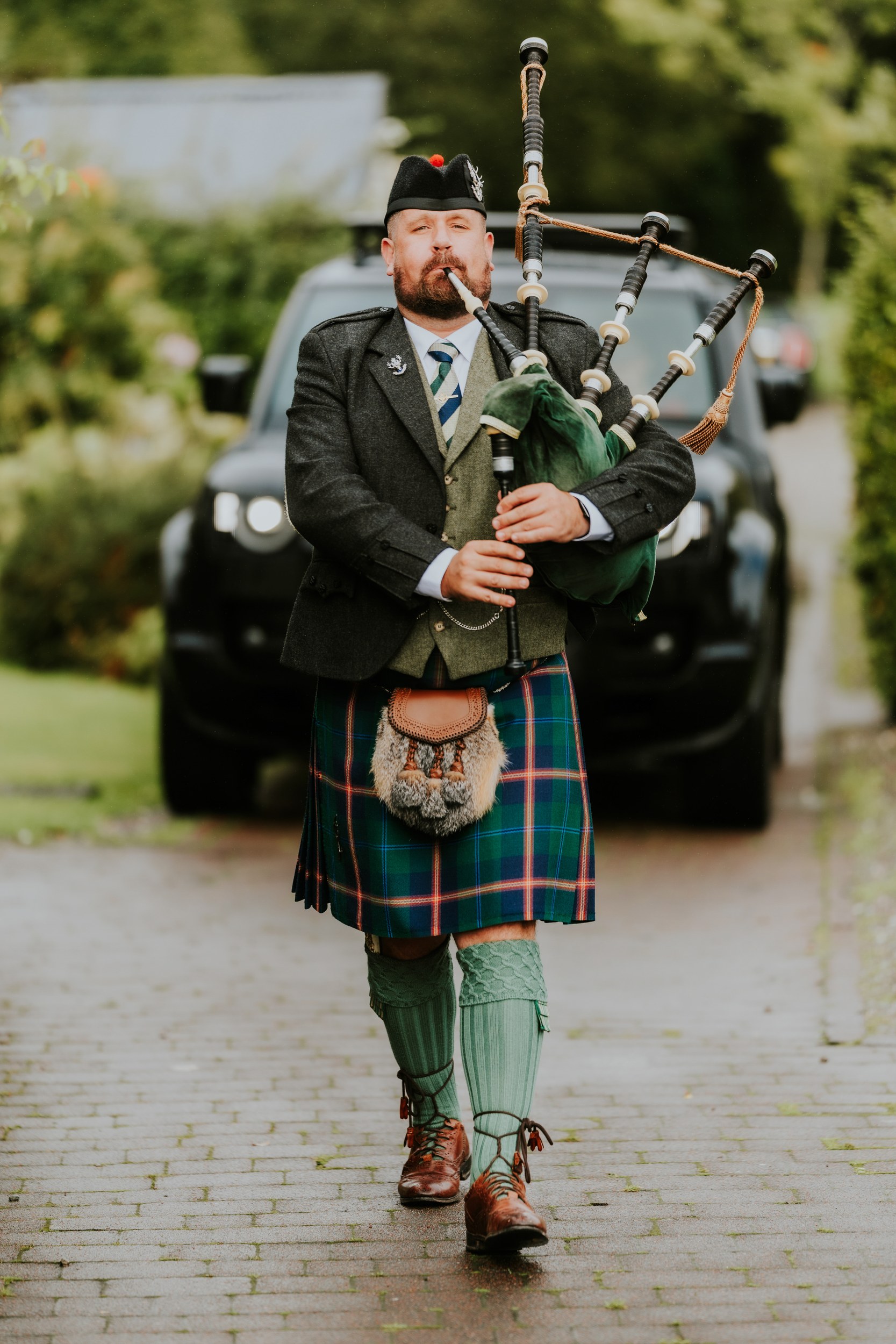 Piper in Highland dress playing bagpipes while walking along a wet paved path, car and trees blurred behind him.