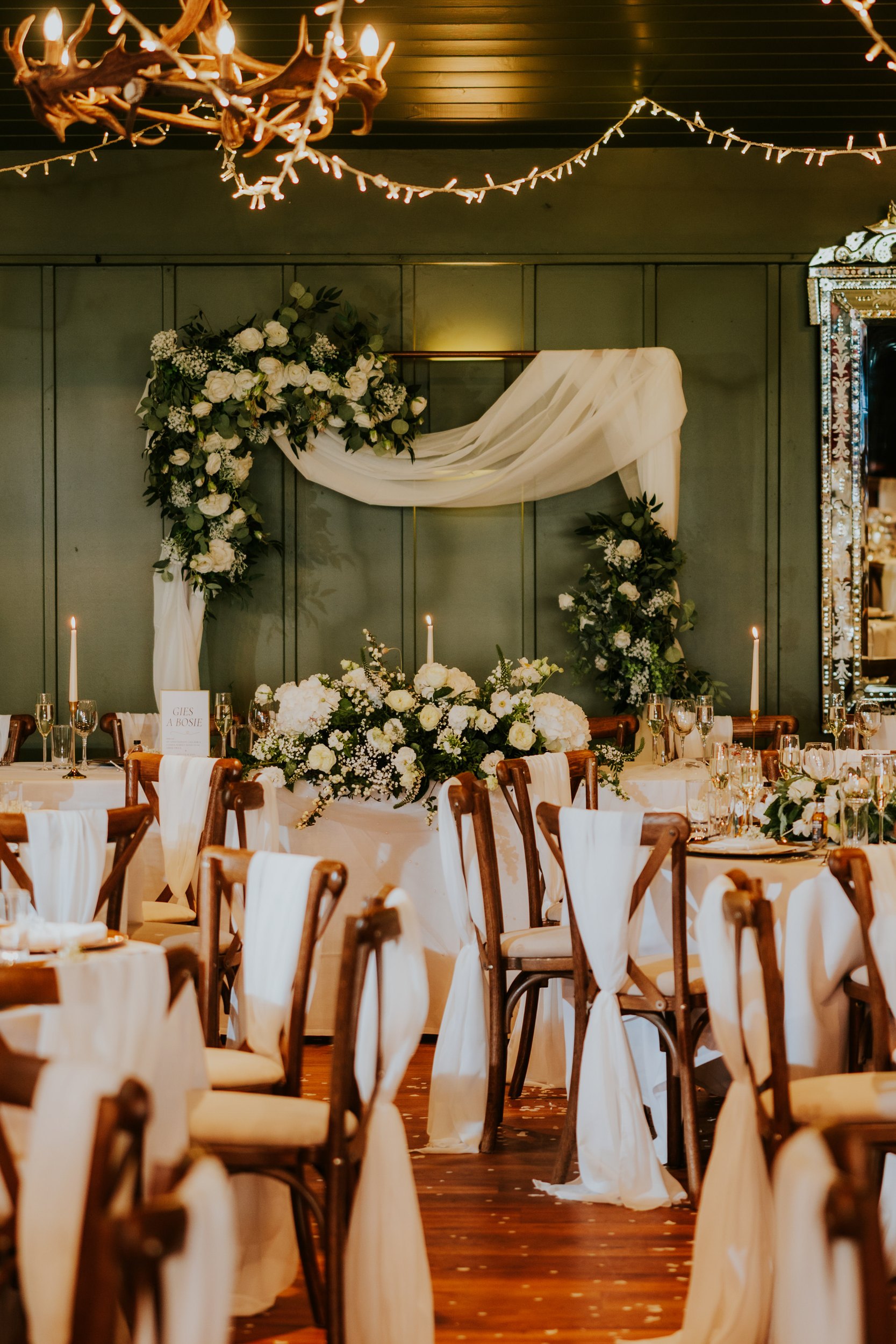 Reception head table with floral arch, draped fabric, candles, fairy lights and chairs tied with white sashes.