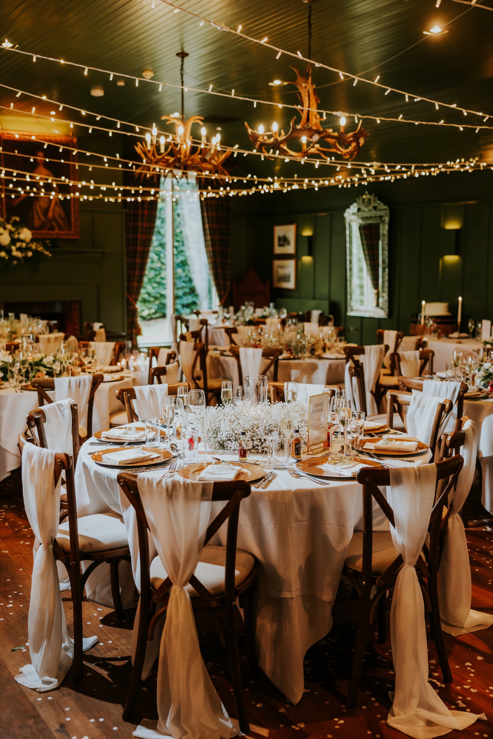 Reception room with round tables set for a wedding, draped chairs, baby’s-breath centrepieces and string lights overhead.