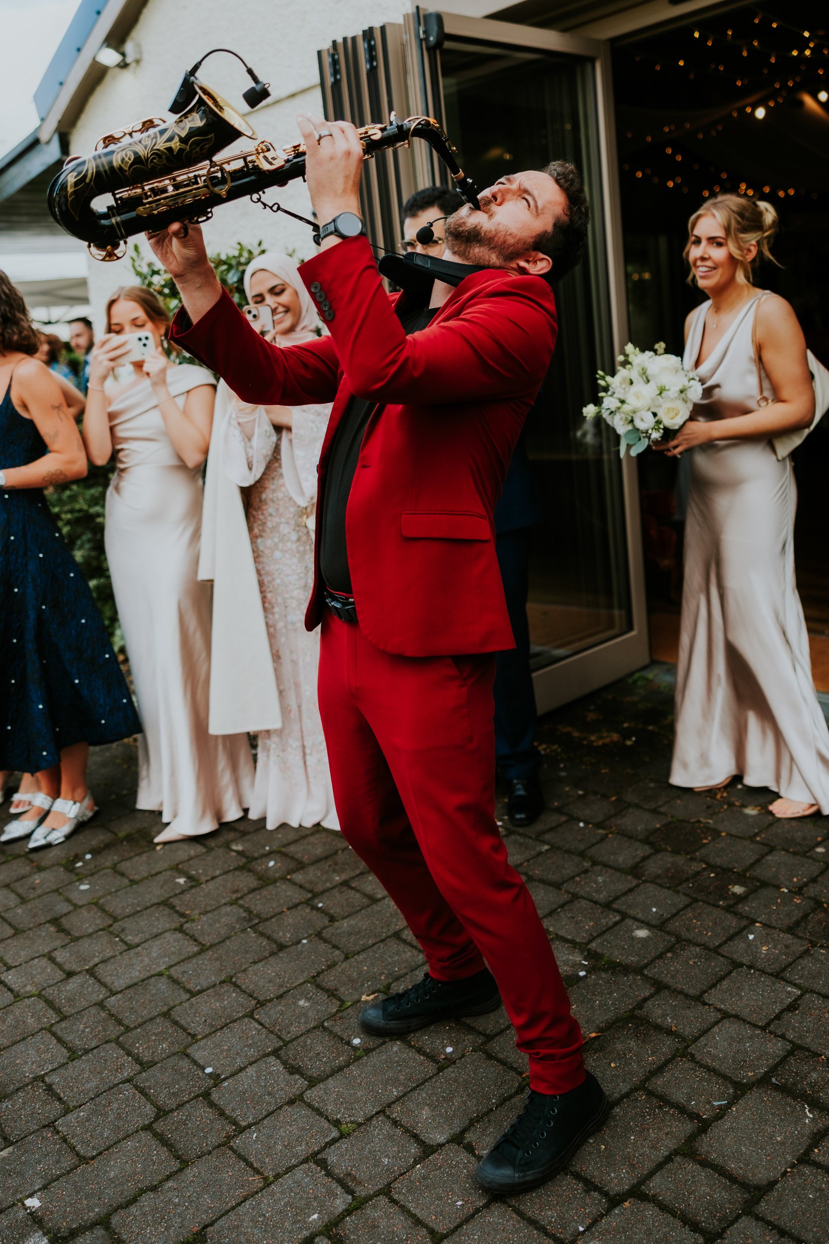 Saxophonist in a red suit playing outdoors as bridesmaids and guests watch and film on phones.