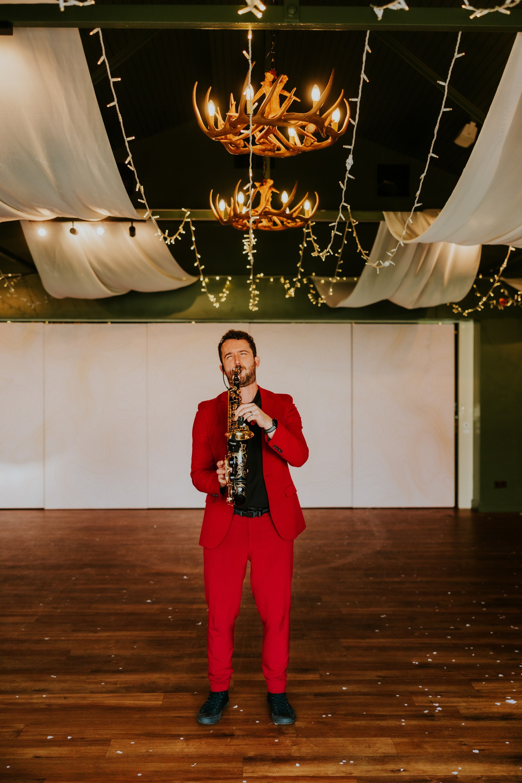 Saxophonist in a red suit playing under antler chandeliers and draped, fairy-lit ceiling in a reception hall.