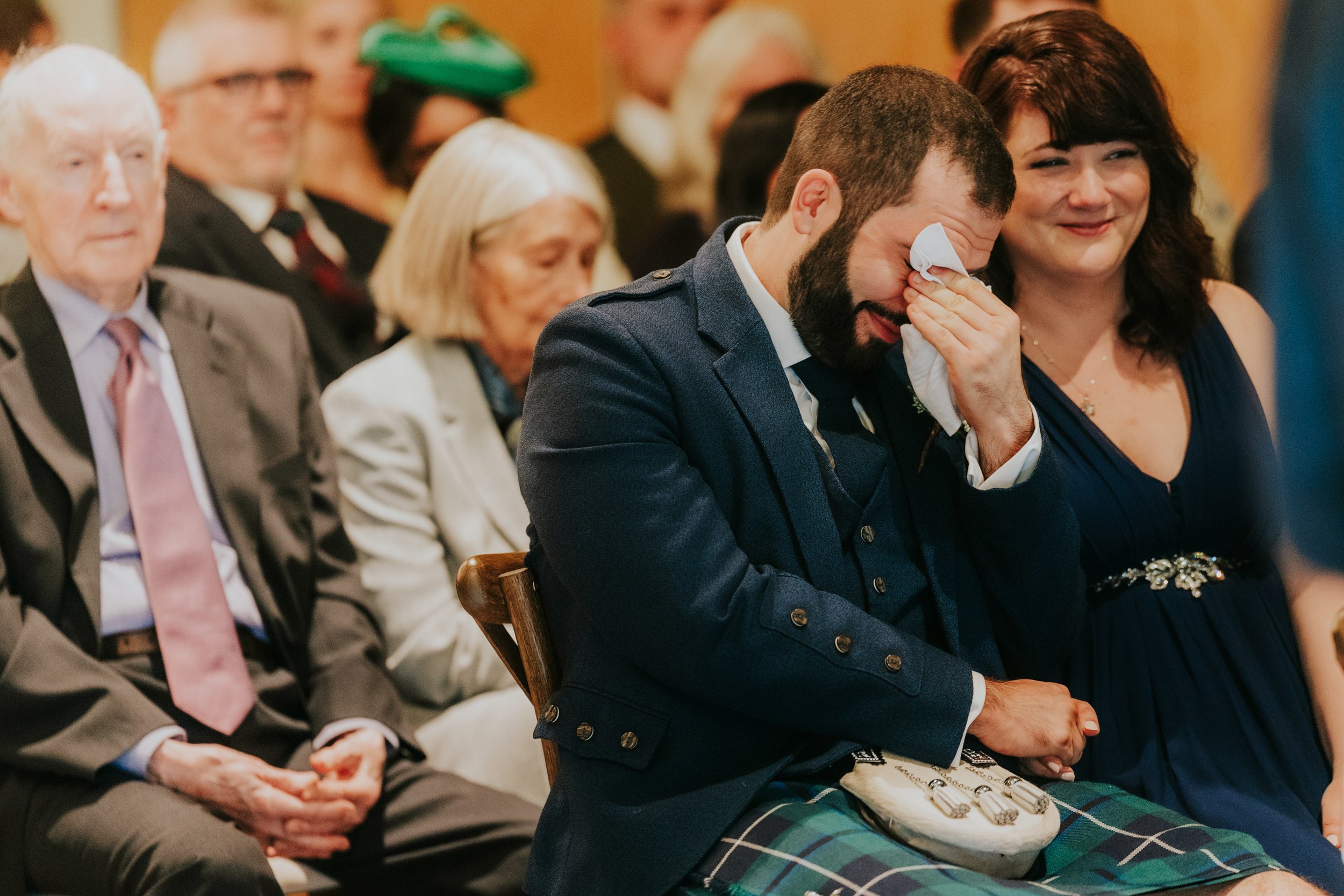 Seated male guest in a kilt wipes his eyes with a tissue while the woman next to him smiles during the ceremony.