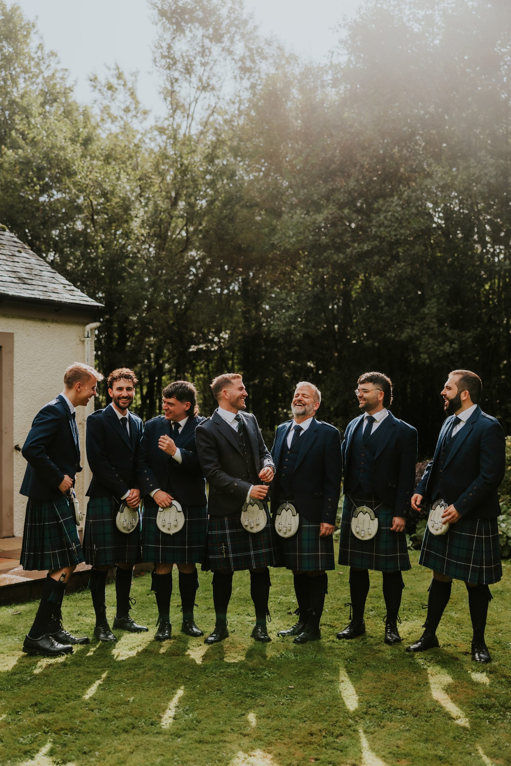Seven men in kilts and jackets standing on grass, smiling and talking outdoors with trees behind them.