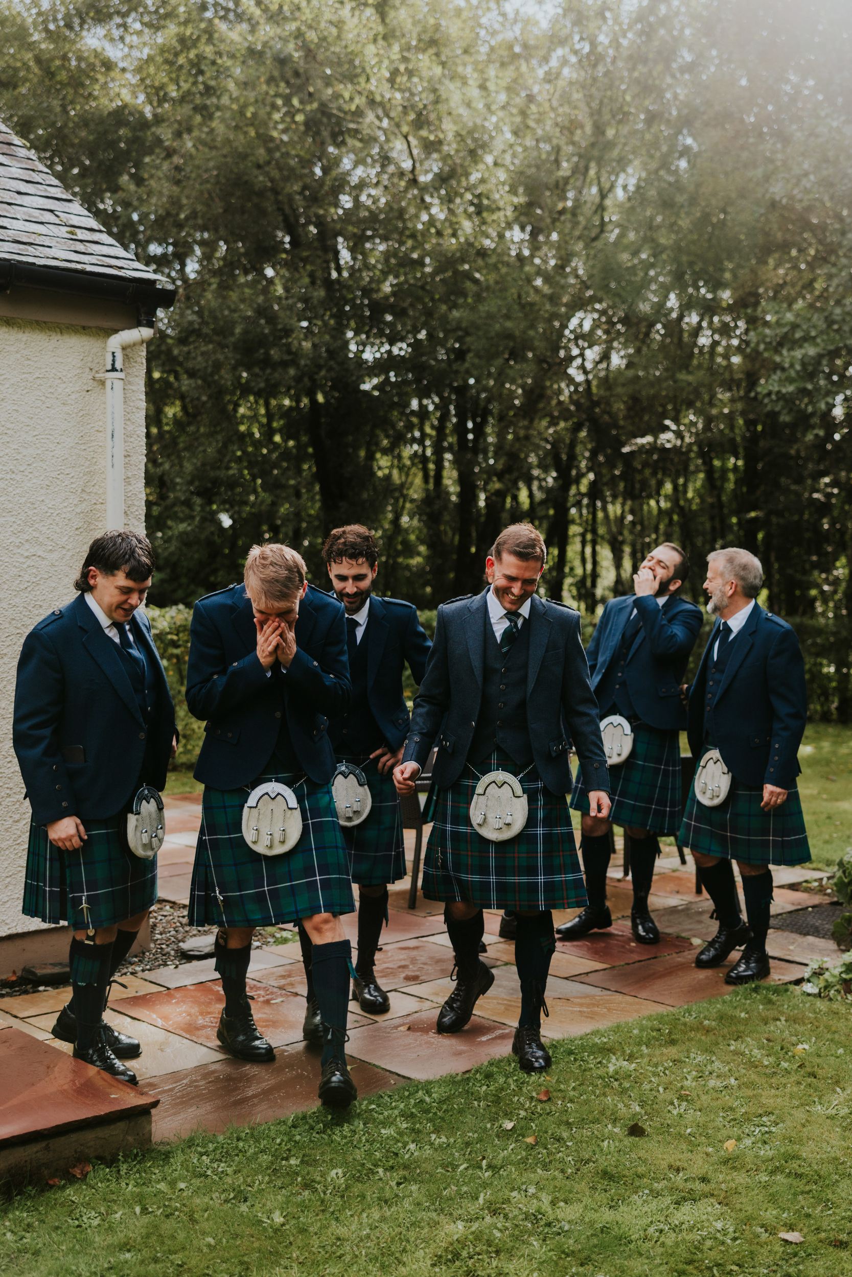 Six men in tartan kilts and jackets walking and laughing outside by a cream building with trees behind.
