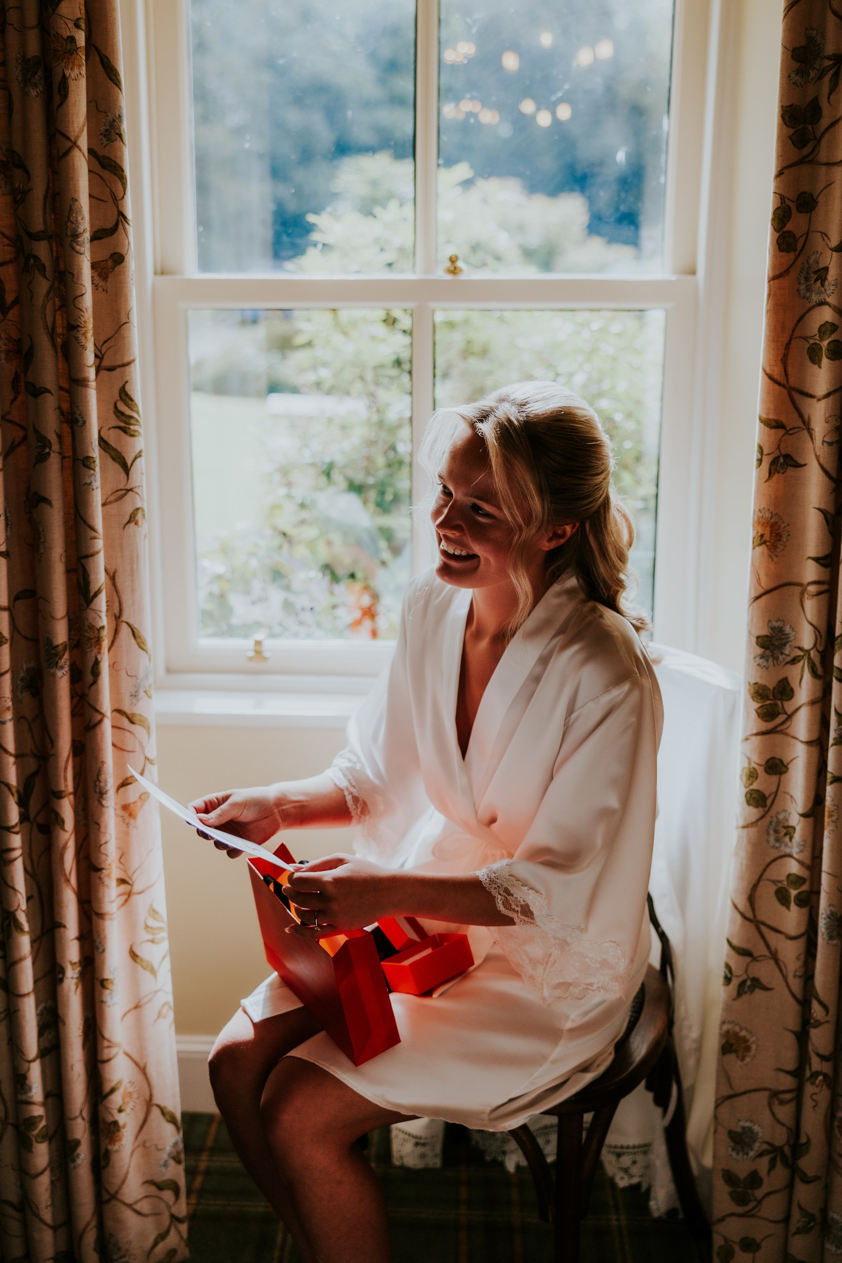 Smiling bride in a white robe sits by a window opening a red gift bag.