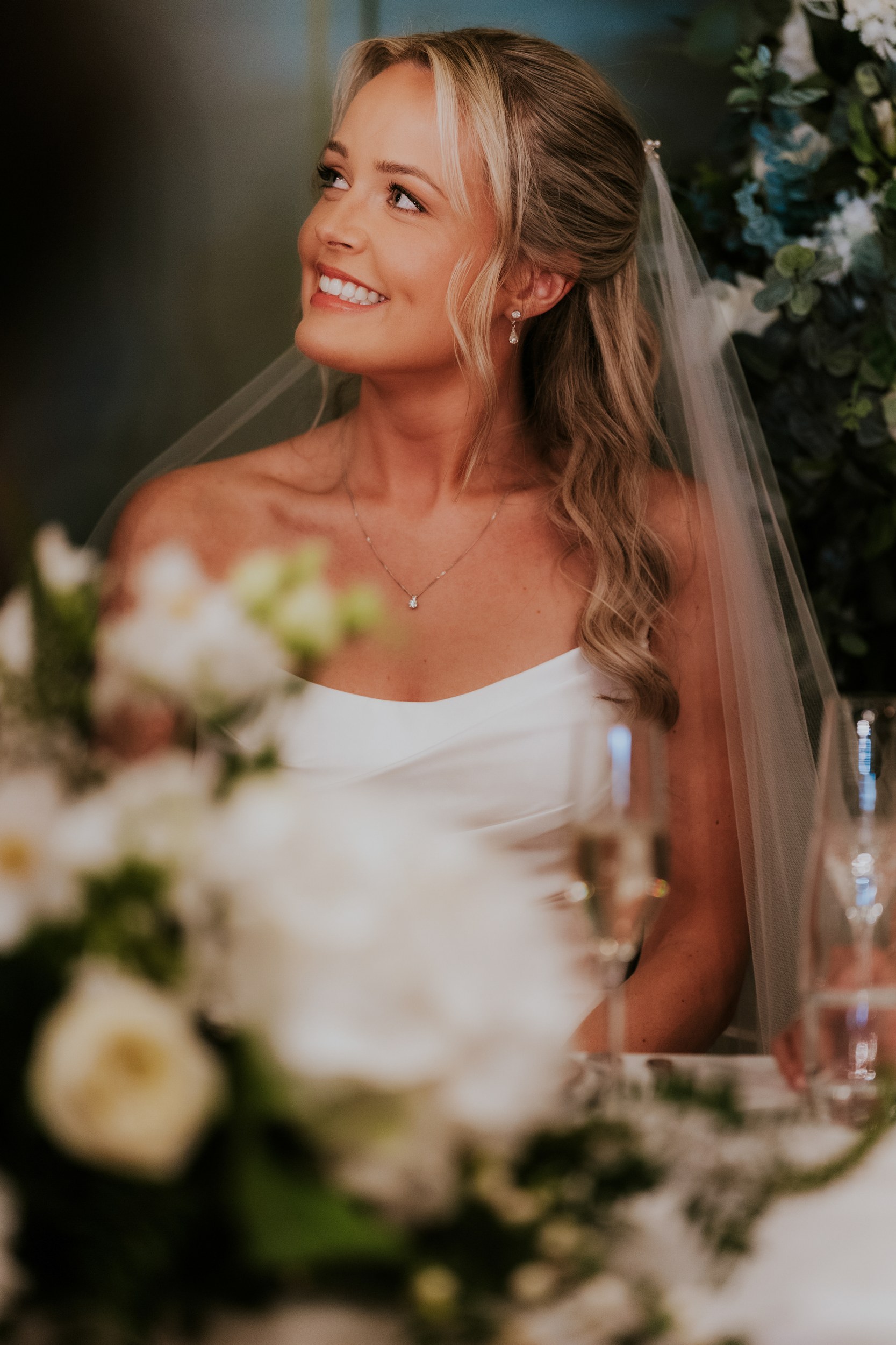 Smiling bride in veil and strapless dress, seated at a table with blurred flowers in the foreground.