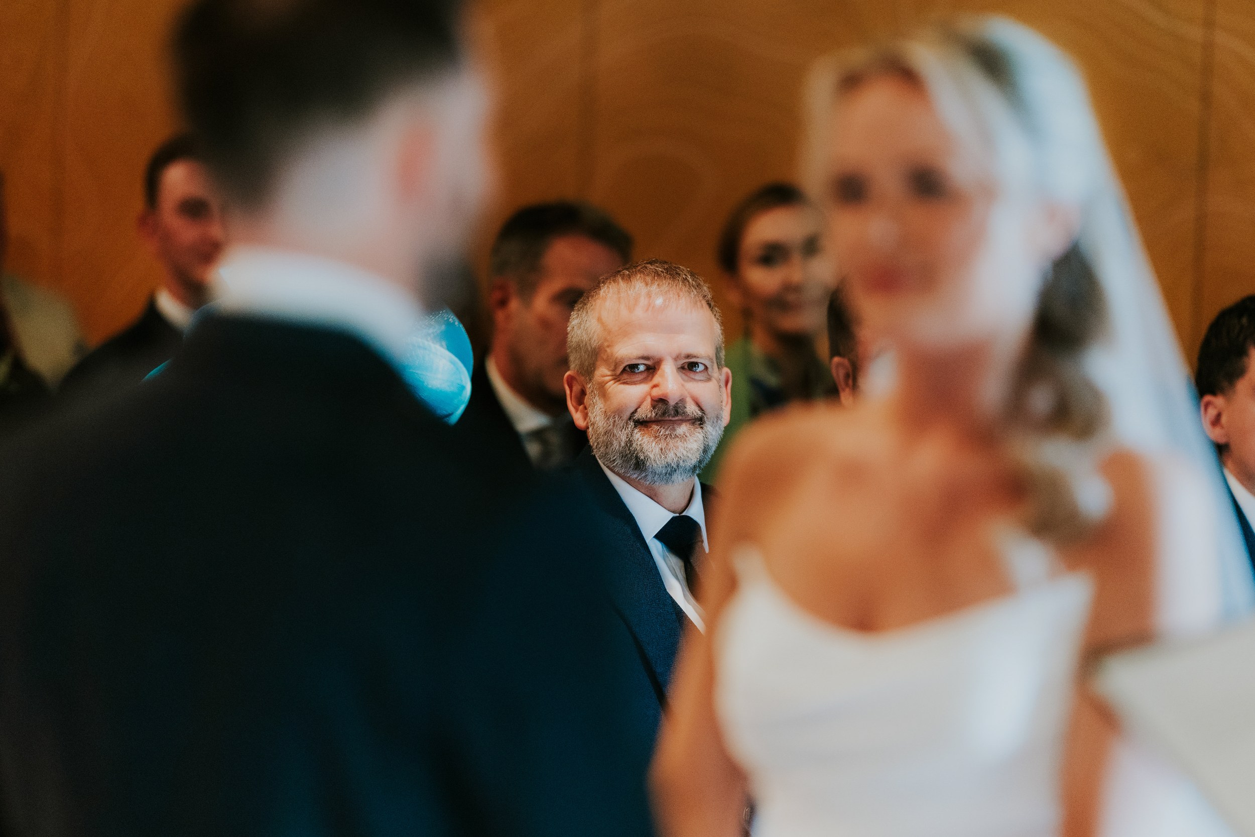 Smiling guest framed between a blurred groom and bride during the ceremony.