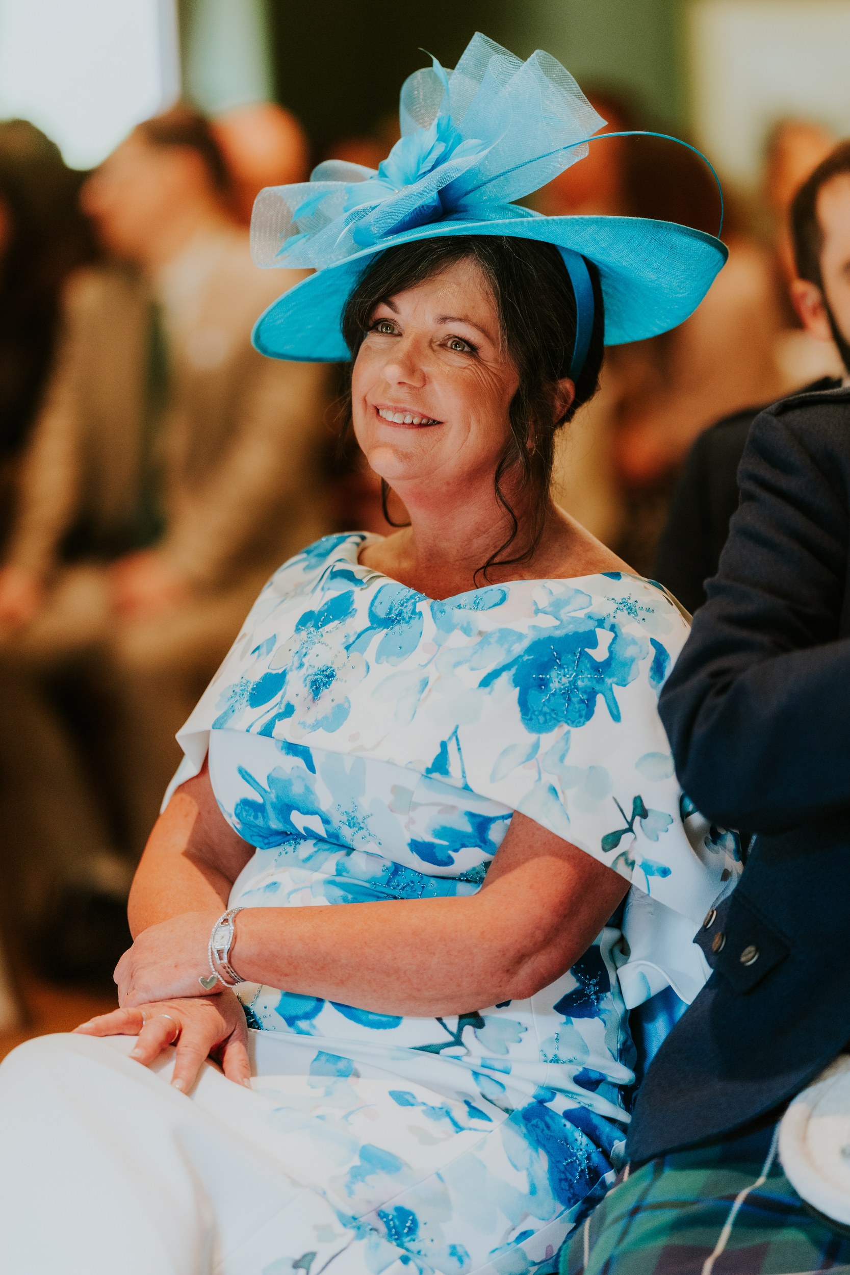 Smiling wedding guest in a turquoise fascinator and blue floral dress, seated among other guests.