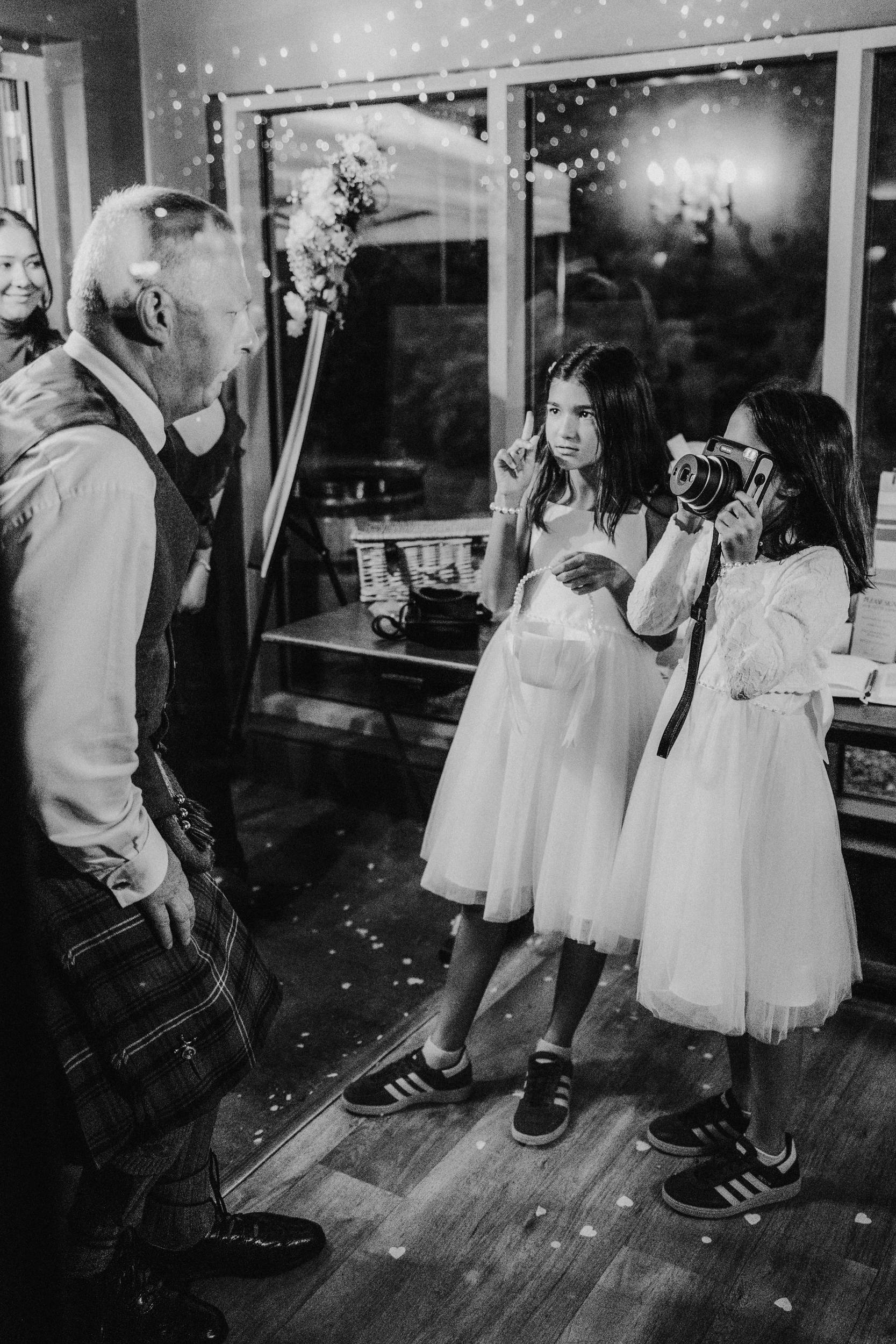 Two flower girls in white dresses, one photographing with an instant camera, the other holding a basket and gesturing to a man in a kilt.