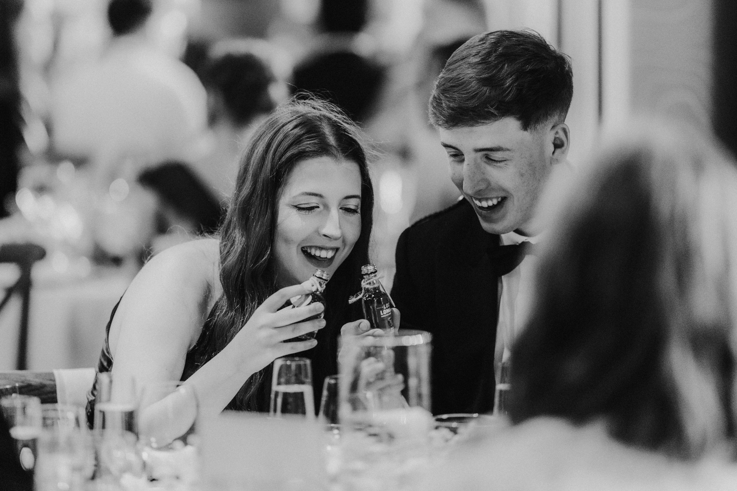 Two guests at a wedding reception laugh and clink small bottles of whisky at a table.