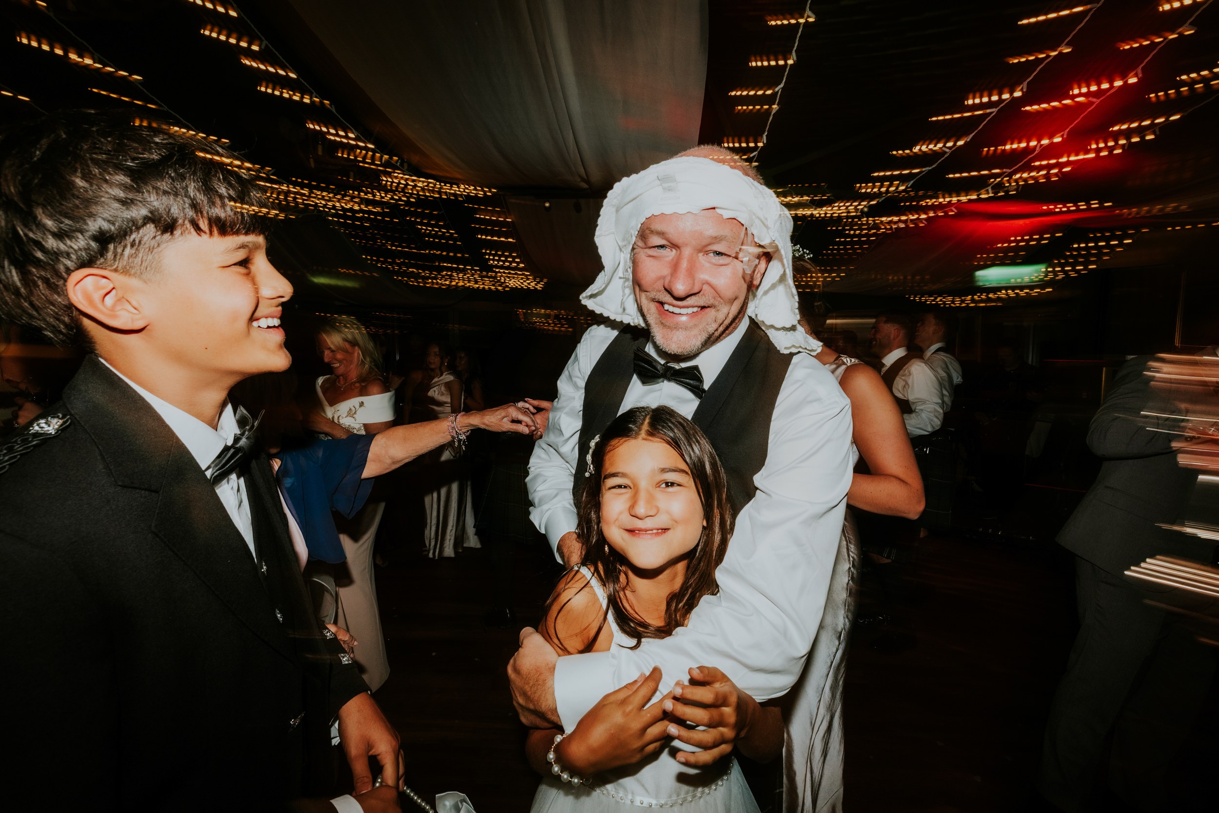 Wedding guests dancing; man with cloth on his head hugs a smiling girl under twinkling string lights.