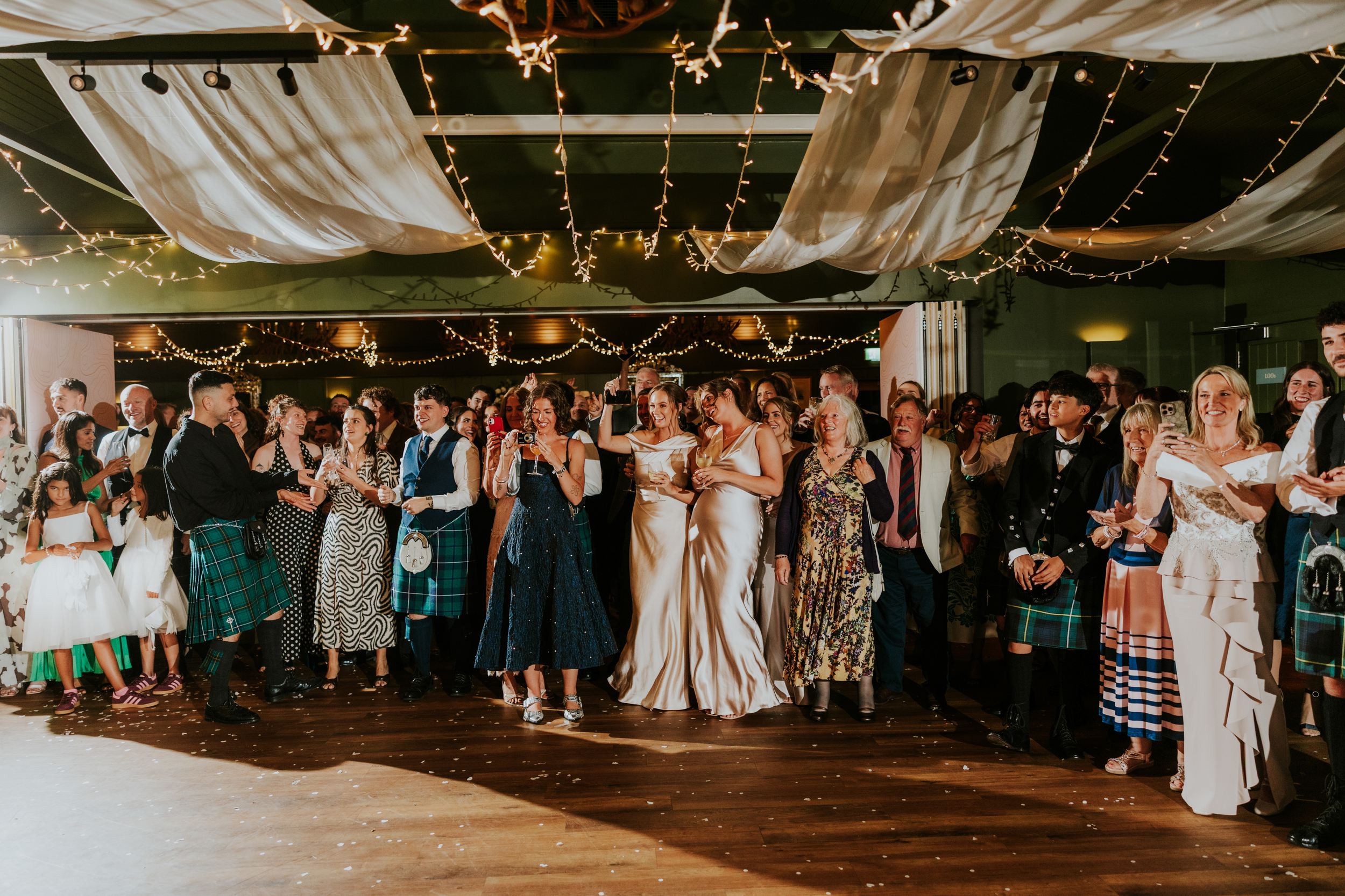 Wedding guests under draped ceiling lights, smiling and taking photos during the reception.