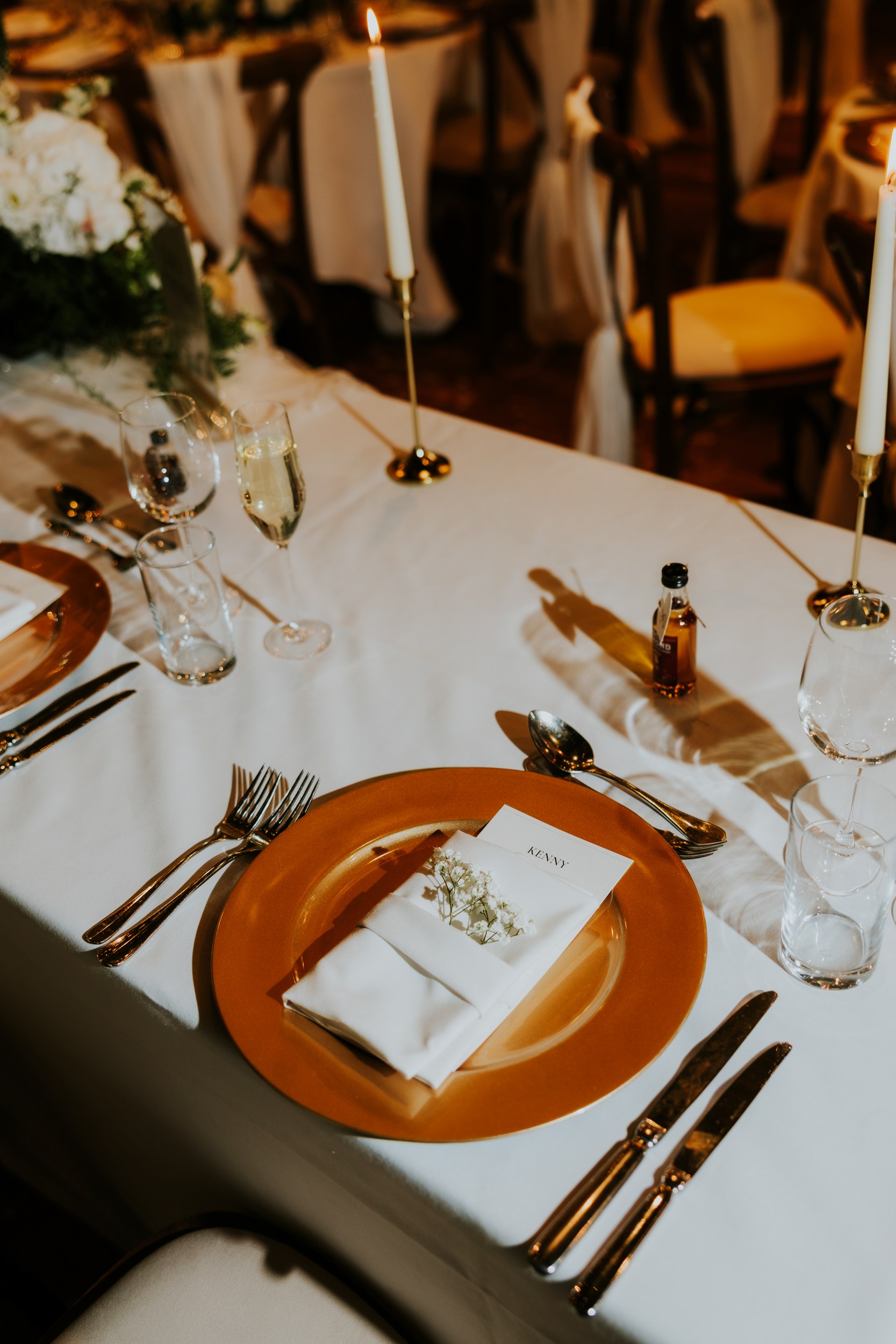 Wedding reception place setting with gold charger, napkin and name card, cutlery, glasses, candles and a small whisky bottle.