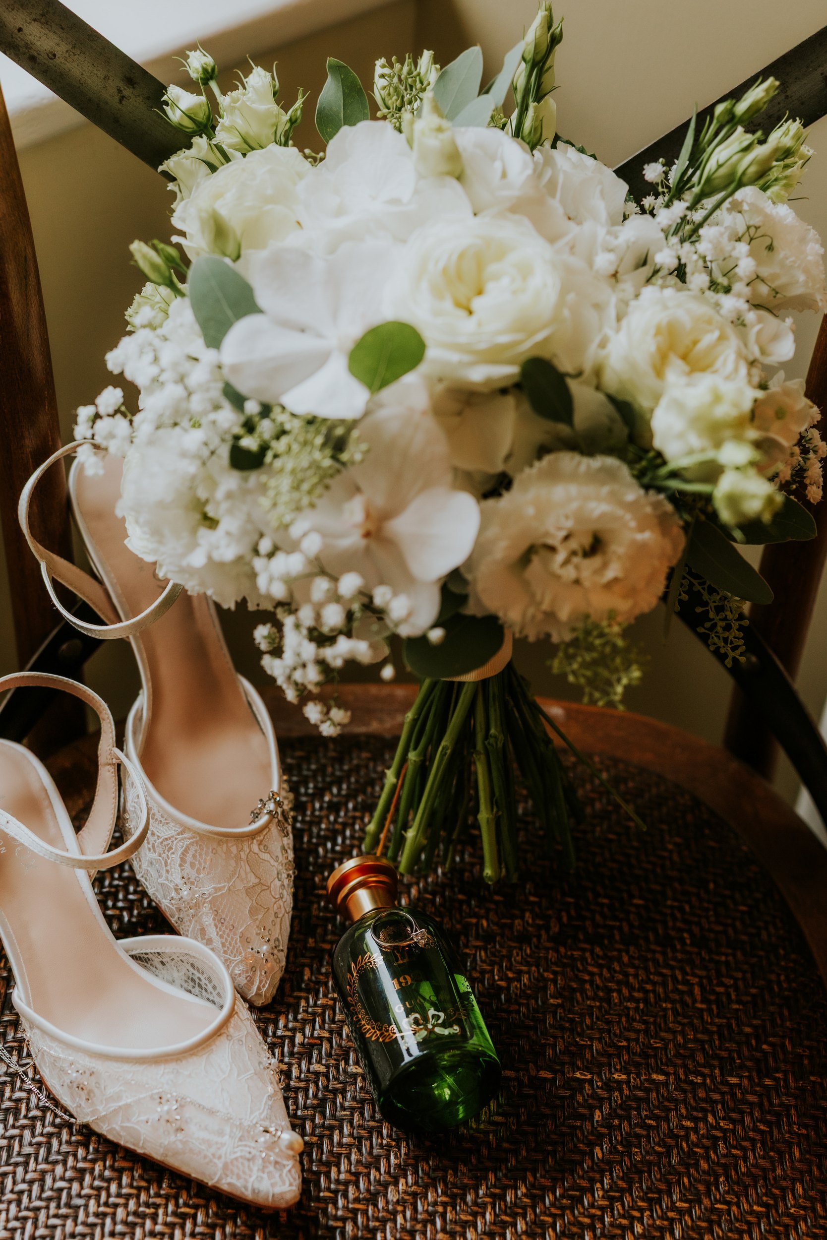 White bridal bouquet on a chair next to lace bridal shoes and a green perfume bottle with rings.