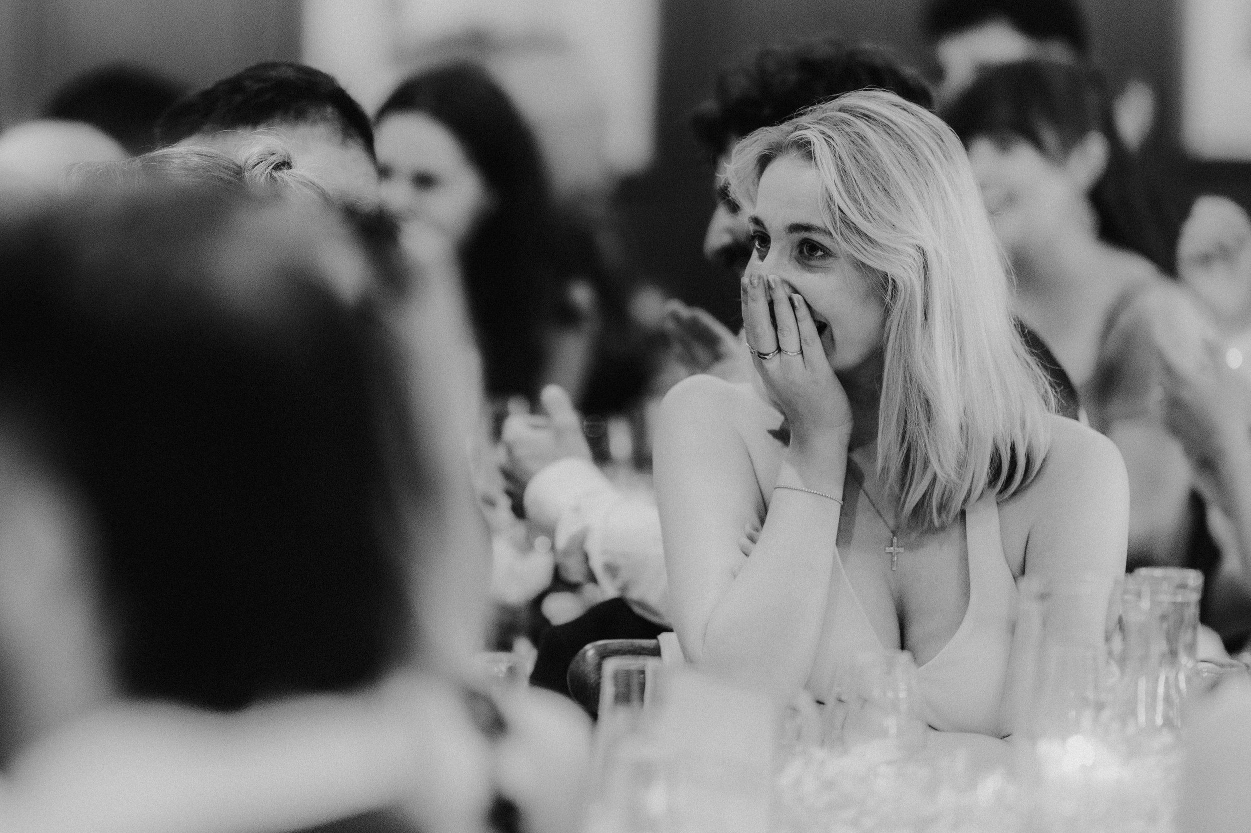 Woman covering her mouth in surprise or emotion while seated at a wedding reception table.
