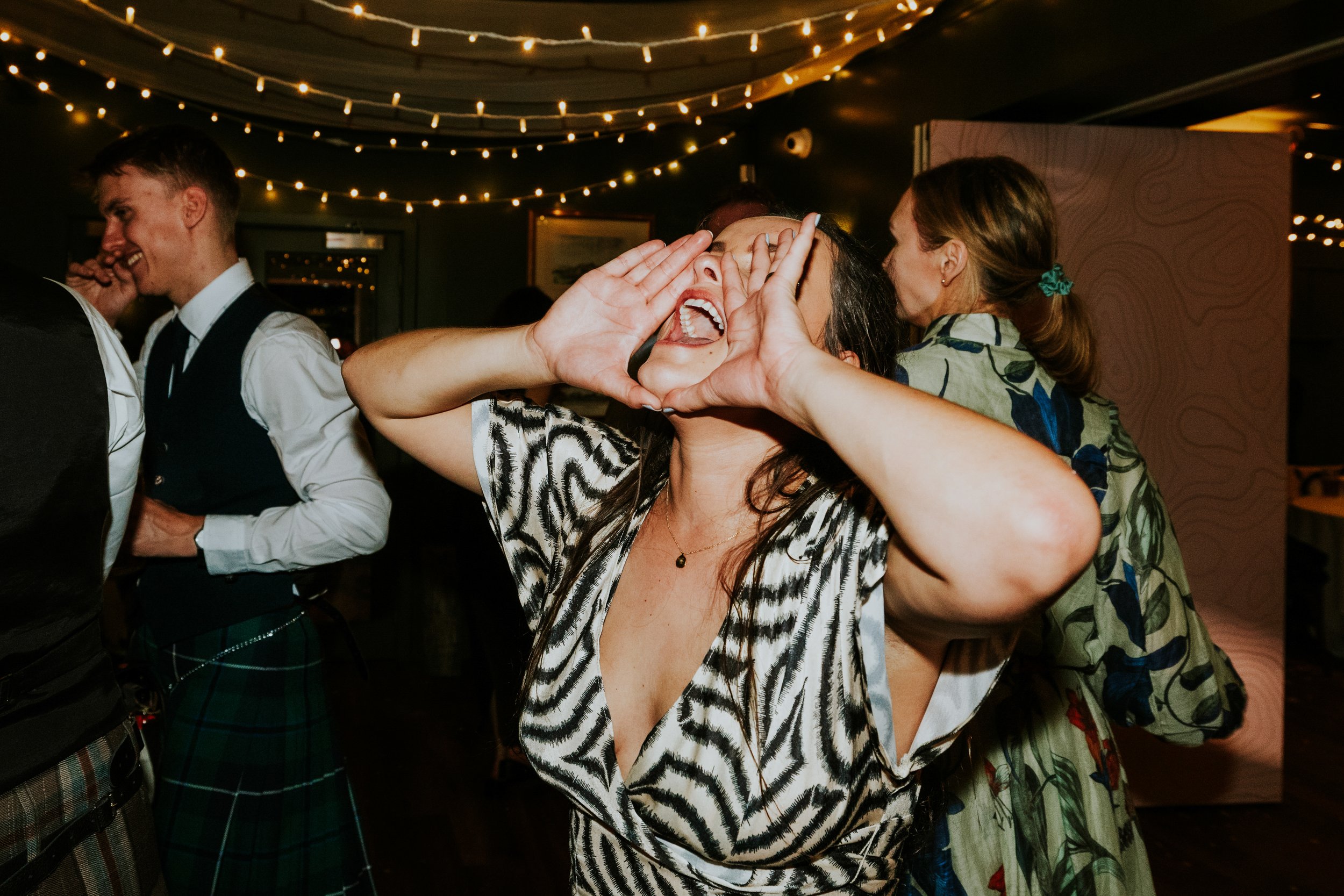Woman cupping her hands and shouting while guests dance under fairy lights at a wedding reception.