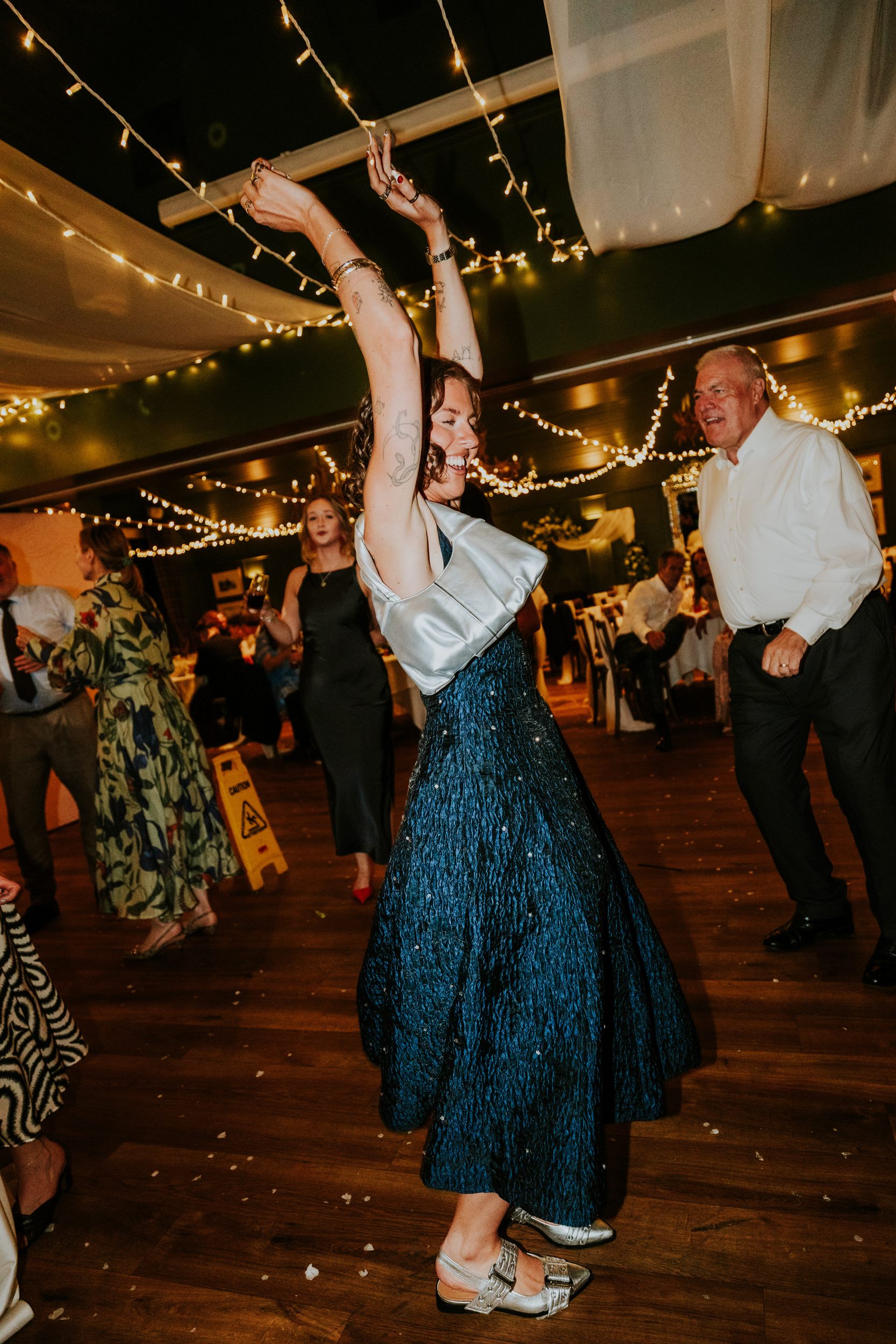 Woman dancing with arms raised under string lights at a wedding reception, with other guests in the background.