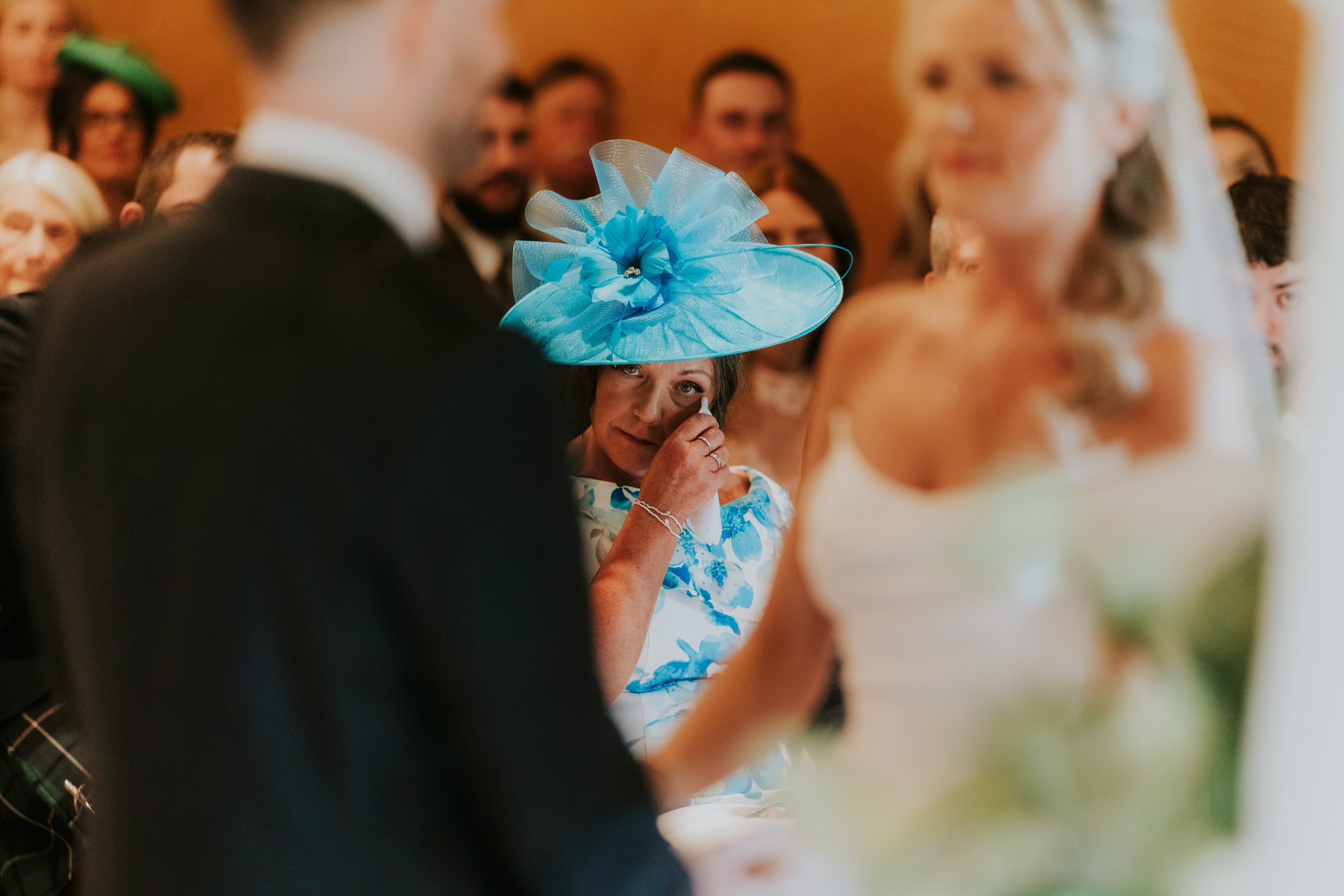 Woman in a turquoise hat wiping a tear as the bride and groom stand blurred in the foreground during the ceremony.