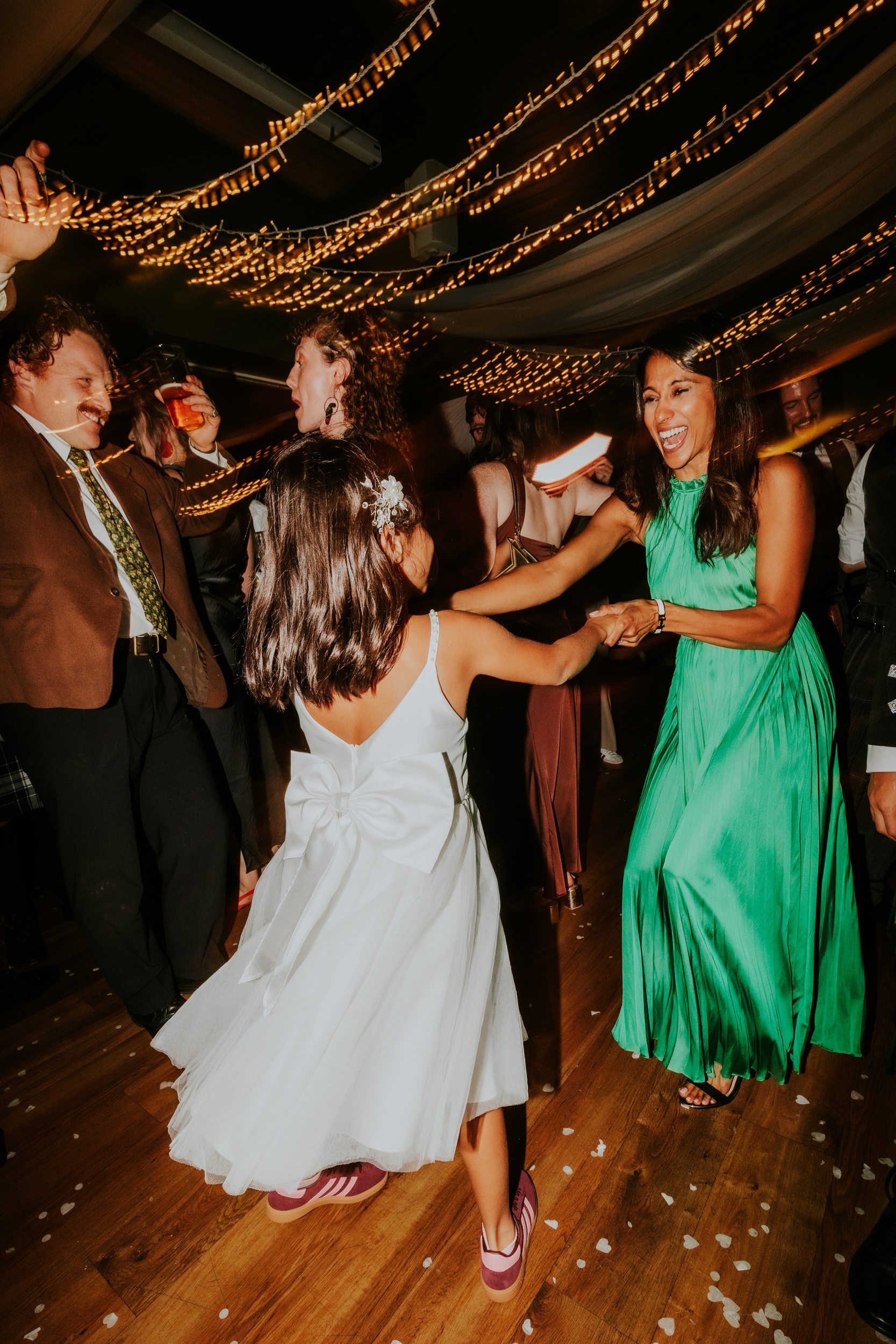 Woman in green dress laughing and dancing with a girl in a white dress under fairy lights.
