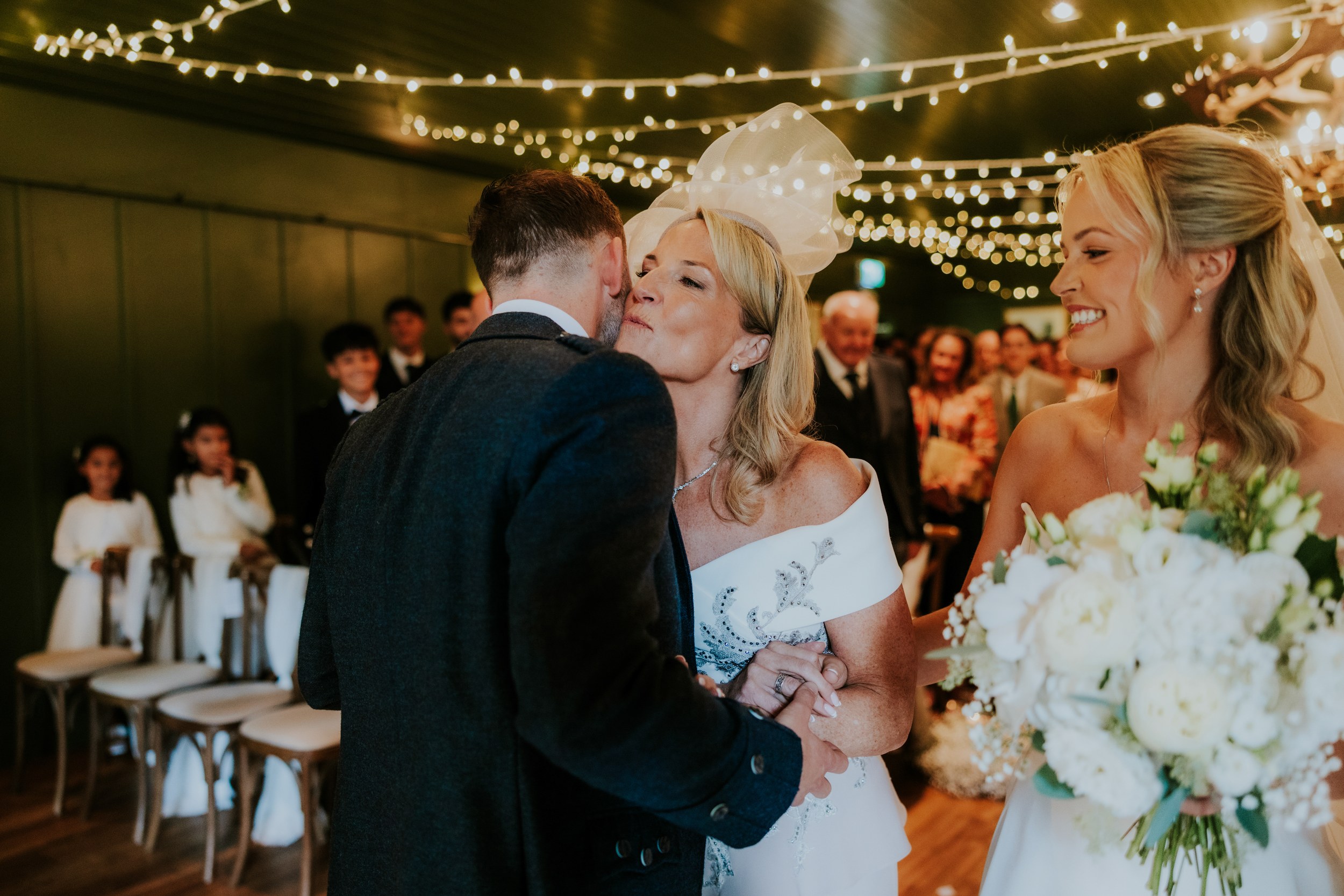 Woman kisses the groom while the bride smiles with her bouquet, guests and flower girls seated under fairy lights.