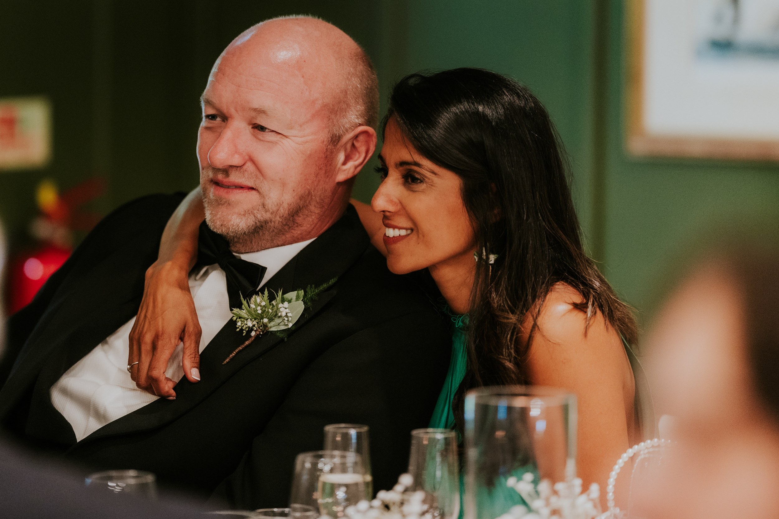 Woman smiling with her arm around a man in a tuxedo at a wedding reception table.
