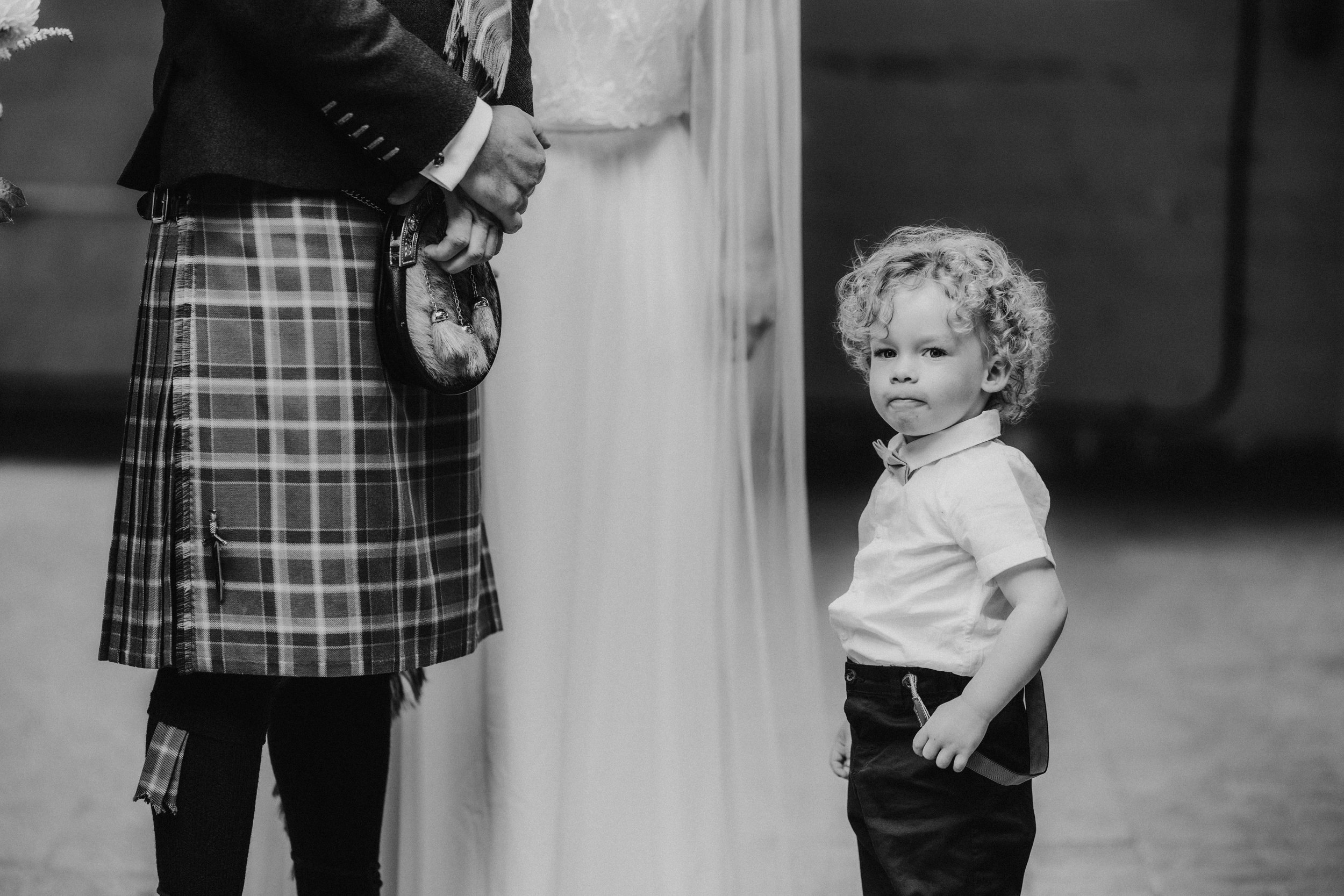 Daniel & Natalie’s Wedding at The Biscuit Factory 18 Black-and-white shot of a person in a tartan kilt beside a wedding dress and a curly-haired child.