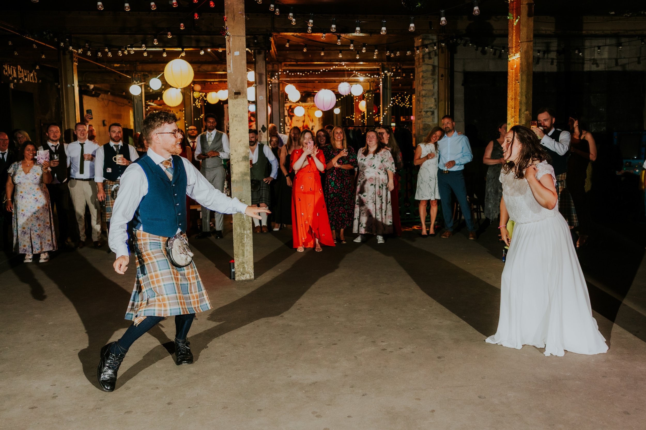 Daniel & Natalie’s Wedding at The Biscuit Factory 82 Bride and groom dancing in an industrial reception space with string lights and guests watching.