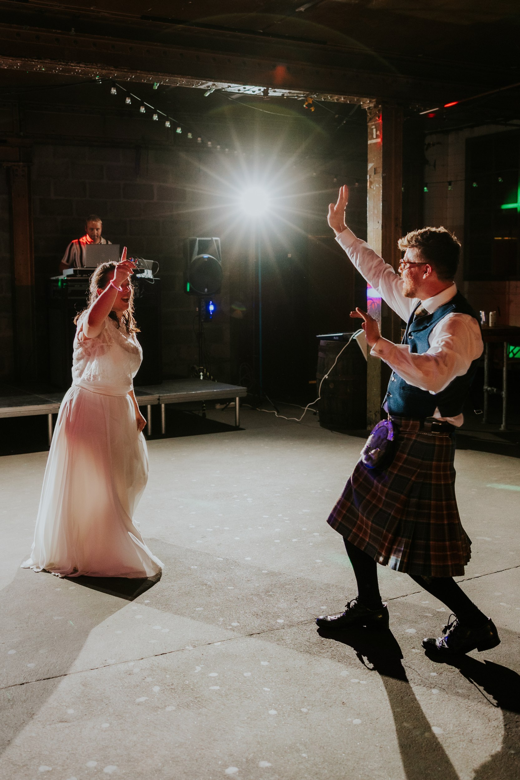 Daniel & Natalie’s Wedding at The Biscuit Factory 81 Bride and groom dancing under a bright backlight in an industrial wedding venue; groom in kilt and bride in white dress.
