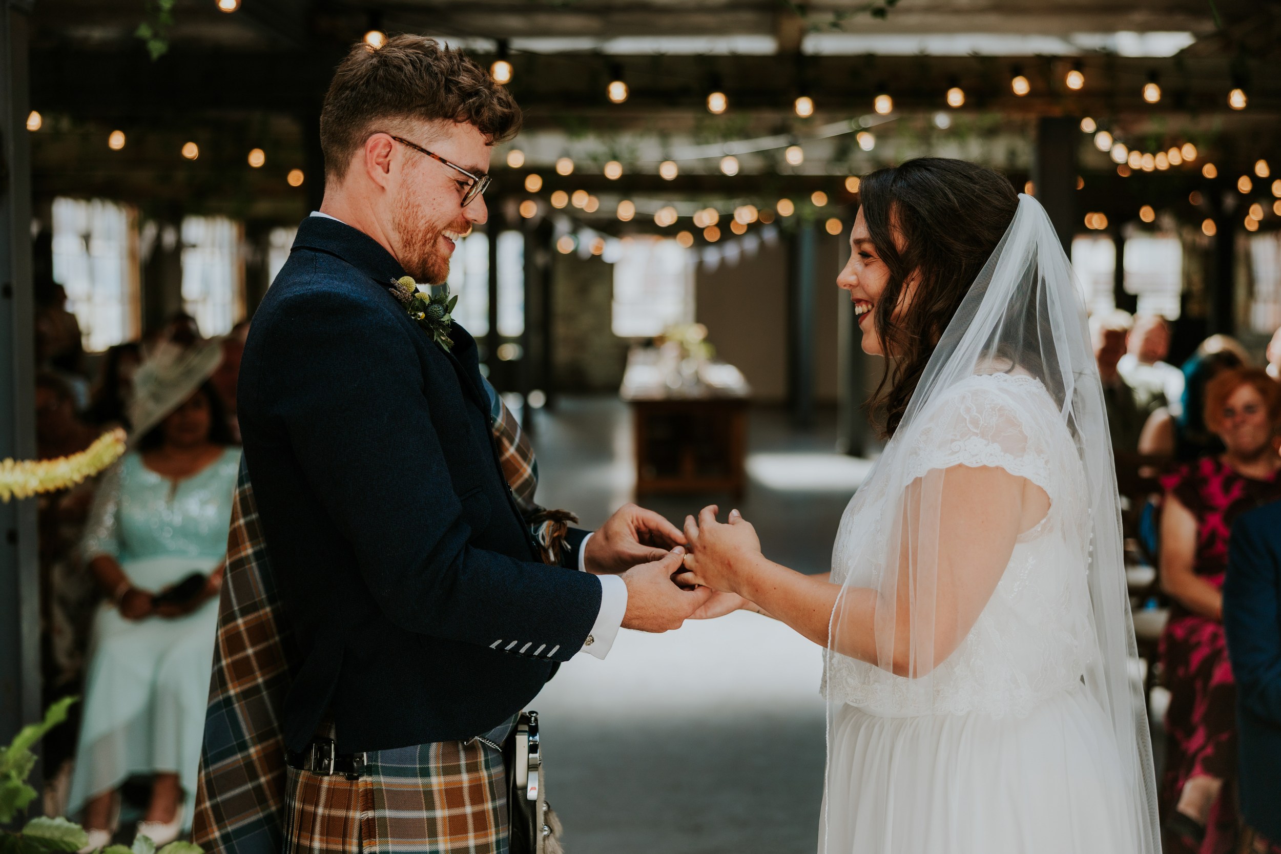 Daniel & Natalie’s Wedding at The Biscuit Factory 26 Bride and groom exchange rings indoors, groom in tartan kilt and bride in lace dress with veil under string lights.