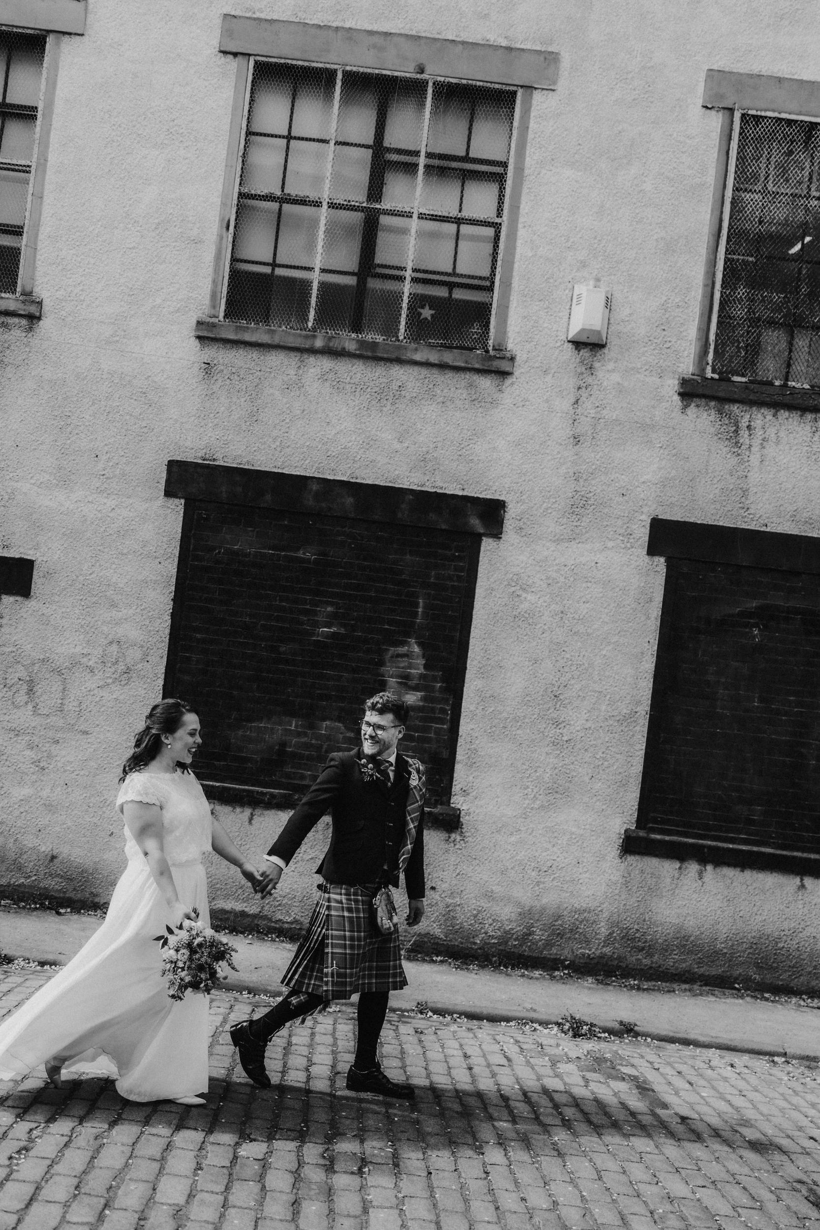 Daniel & Natalie’s Wedding at The Biscuit Factory 44 Bride and groom holding hands, groom in a kilt, walking past an industrial building on a cobbled street.
