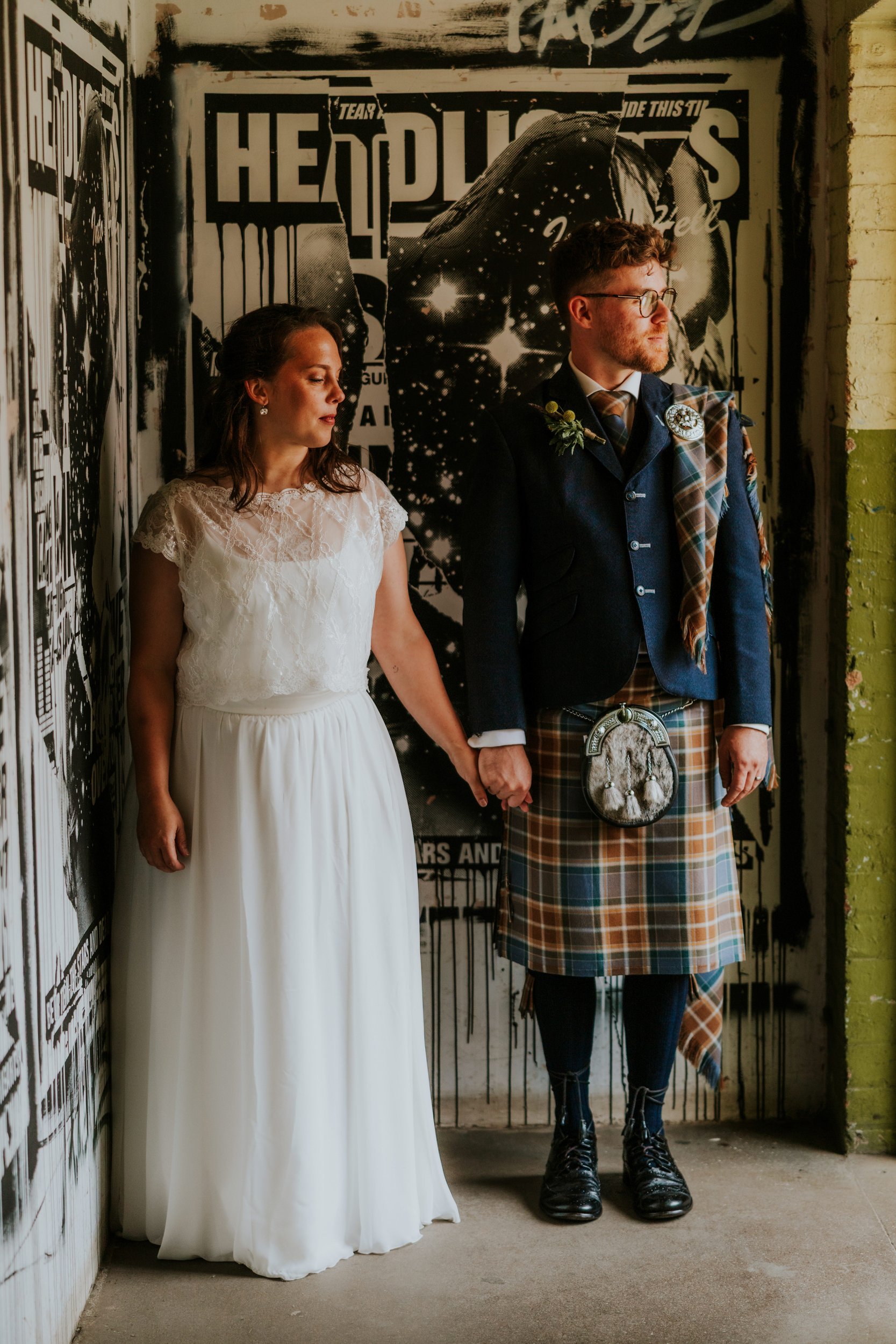 Daniel & Natalie’s Wedding at The Biscuit Factory 50 Bride and groom holding hands in wedding dress and tartan kilt against a black-and-white postered wall.