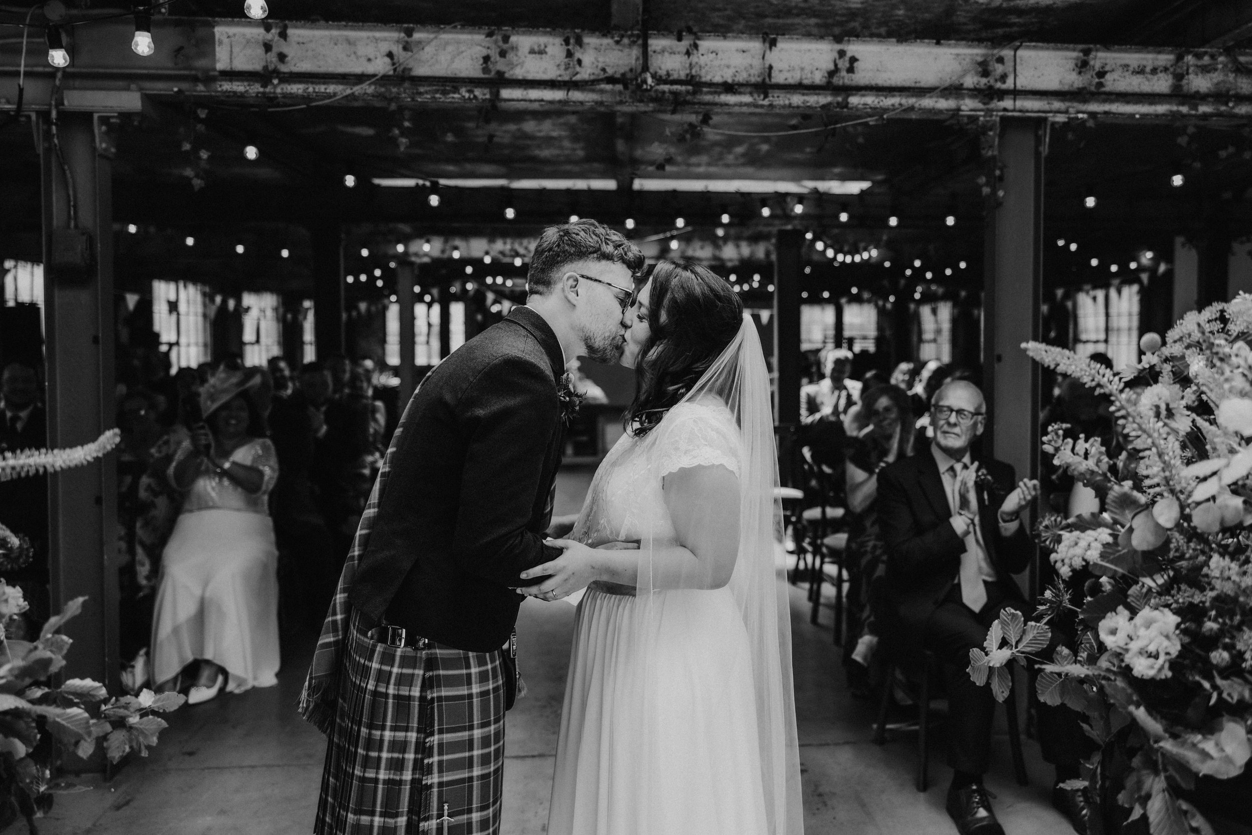 Daniel & Natalie’s Wedding at The Biscuit Factory 27 Bride and groom kiss at ceremony in industrial room with string lights and applauding guests