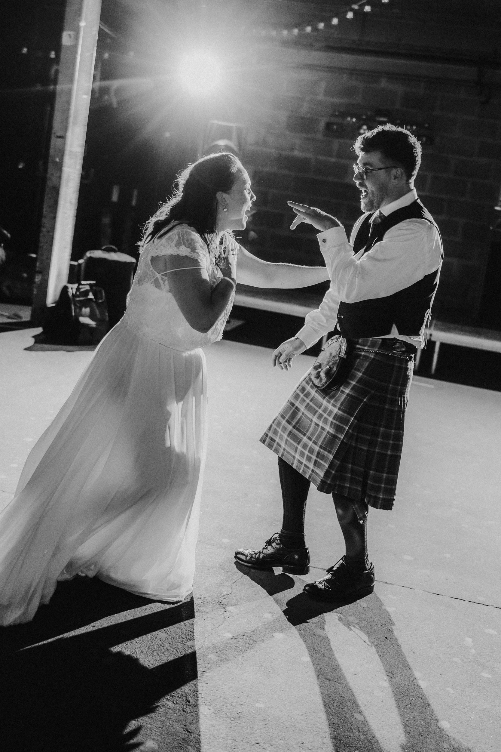 Daniel & Natalie’s Wedding at The Biscuit Factory 83 Bride and groom laughing on the dancefloor; groom in a kilt, bride in a lace-sleeved dress, strong backlight and long shadows.