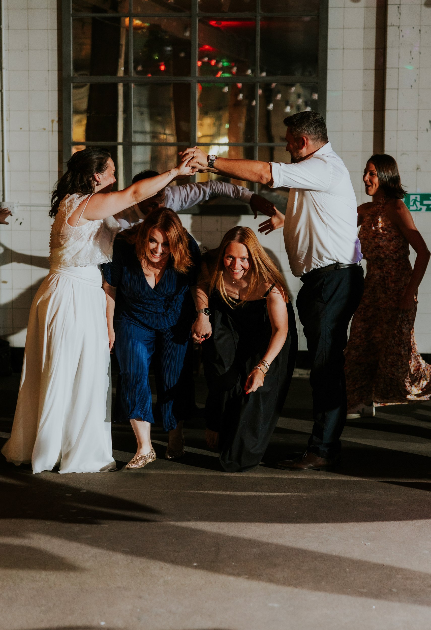 Daniel & Natalie’s Wedding at The Biscuit Factory 77 Bride and guests laughing as they crouch through a hand-arch while dancing at the wedding reception.