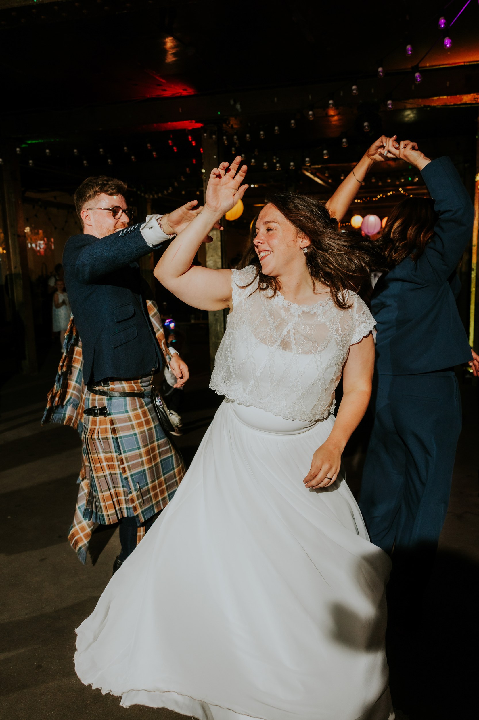 Daniel & Natalie’s Wedding at The Biscuit Factory 73 Bride dancing and smiling at reception with guests under hanging string lights