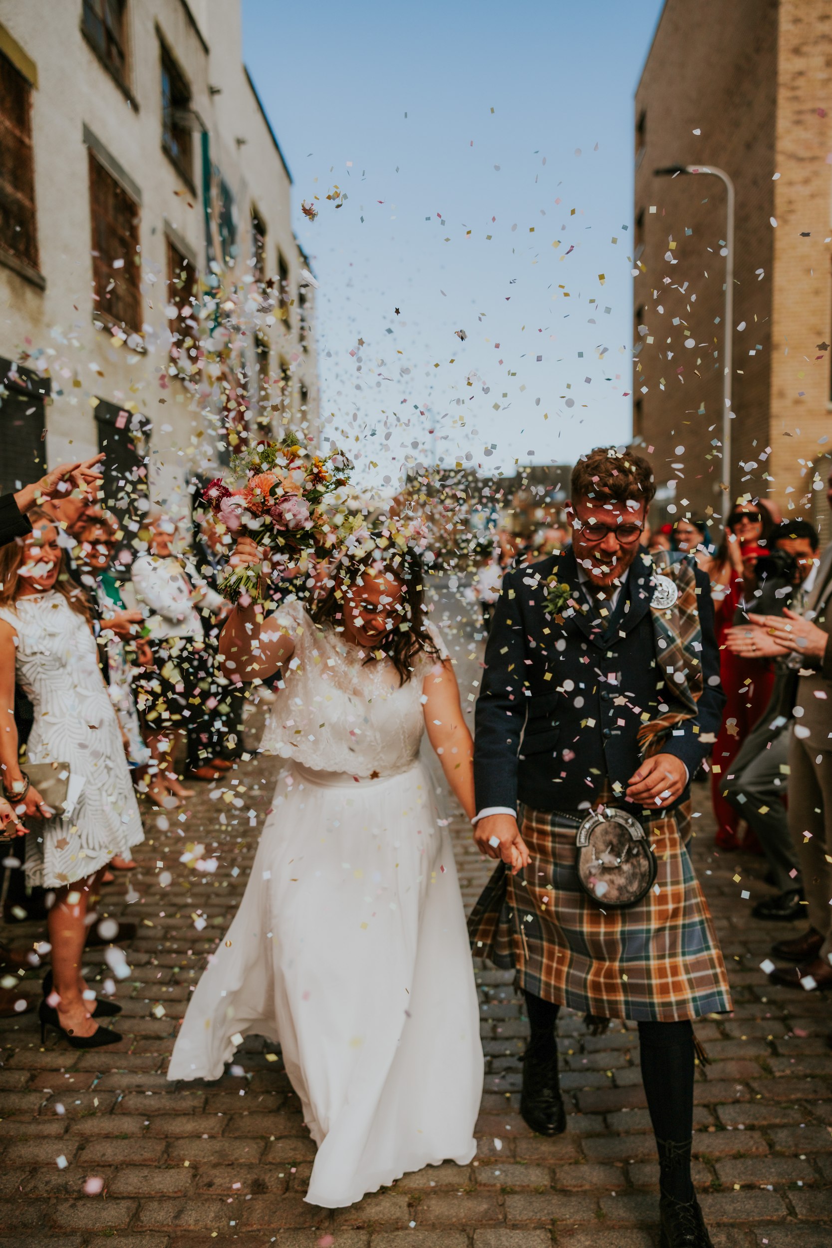 Daniel & Natalie’s Wedding at The Biscuit Factory 34 Bride holding bouquet and groom in tartan kilt walk through falling confetti with cheering guests on a cobbled street.