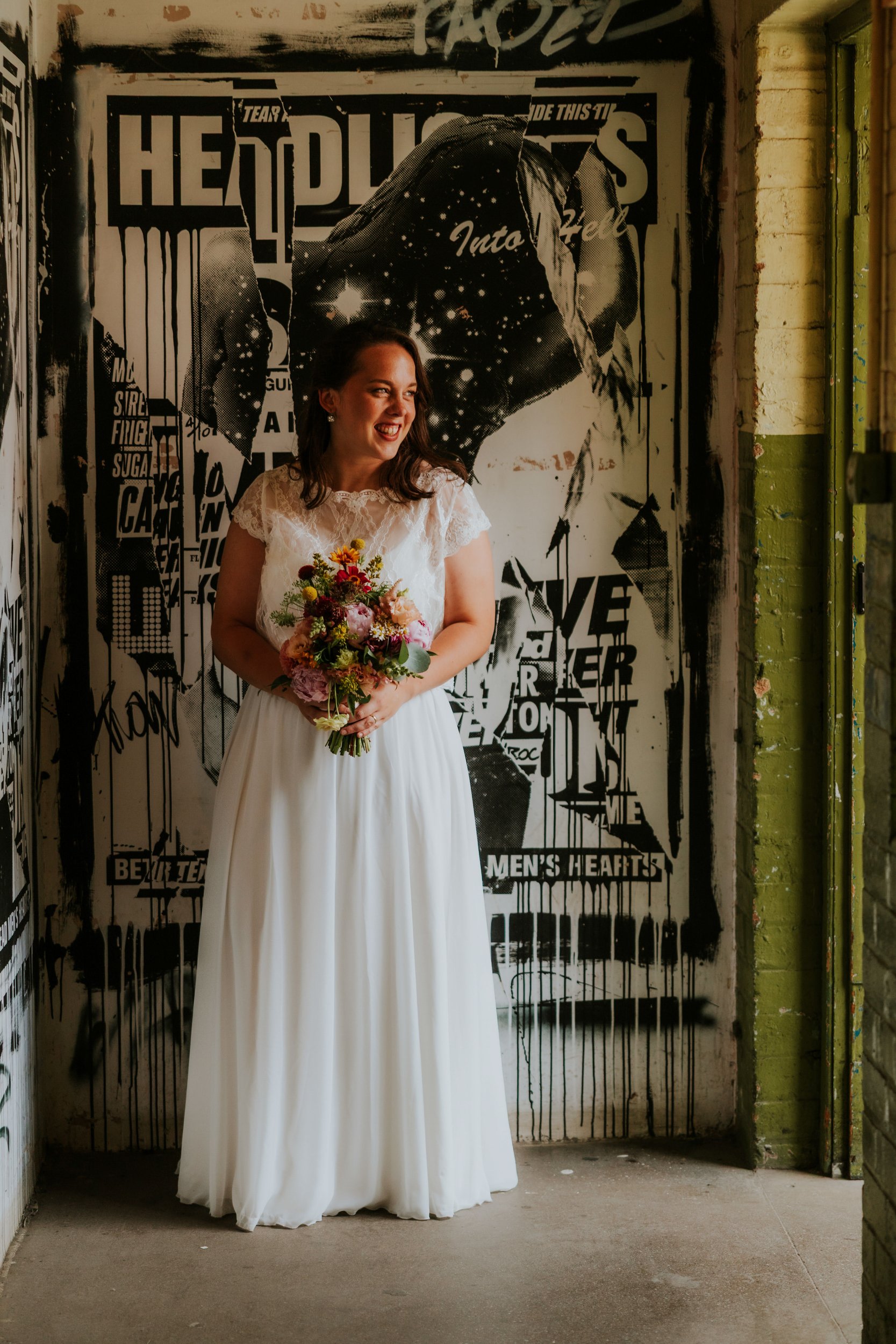 Daniel & Natalie’s Wedding at The Biscuit Factory 48 Bride in lace dress holding bouquet, smiling in front of black-and-white poster at The Biscuit Factory, Edinburgh.
