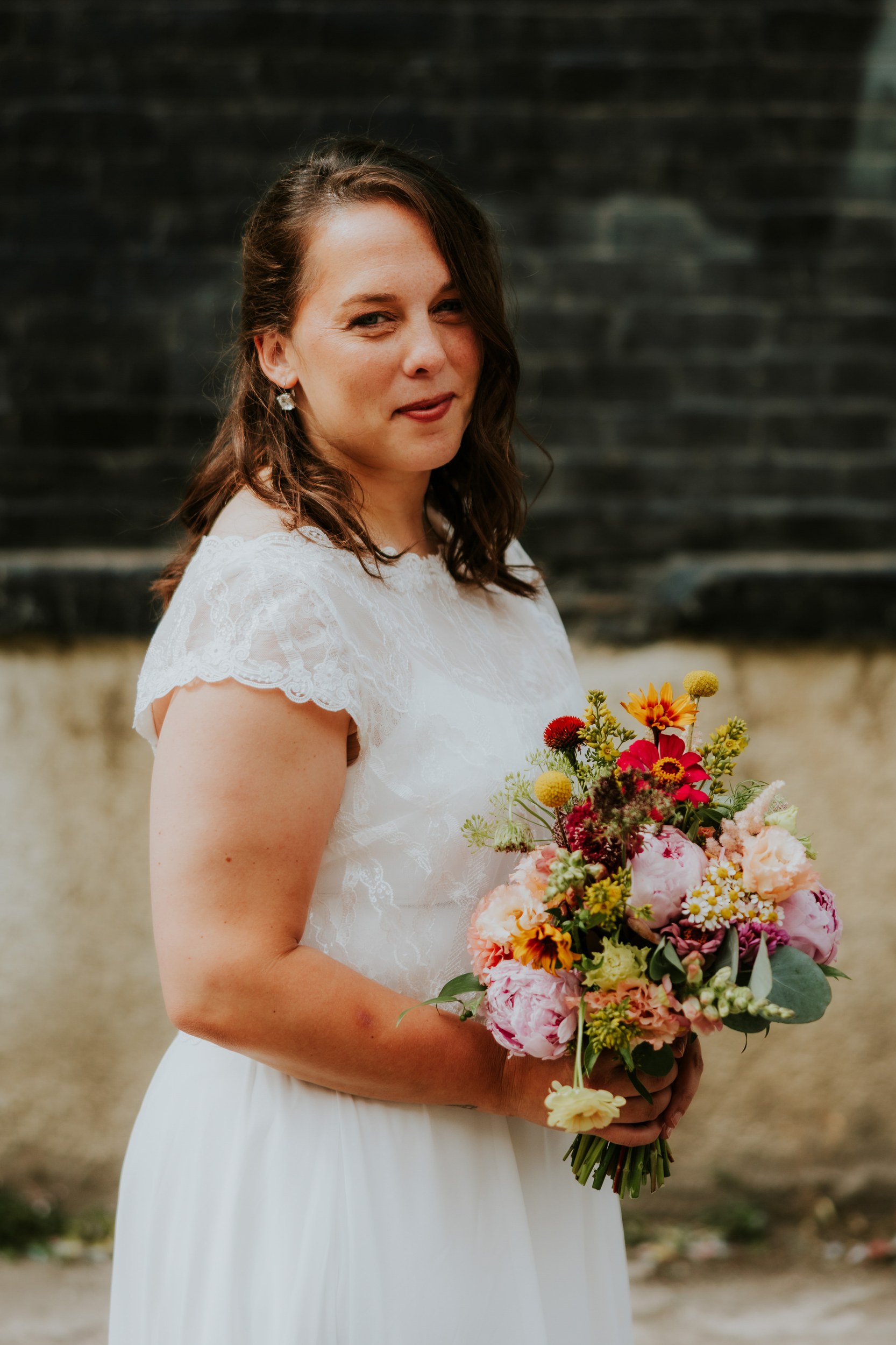 Daniel & Natalie’s Wedding at The Biscuit Factory 43 Bride in lace wedding dress holding a colourful bouquet against a dark brick wall.