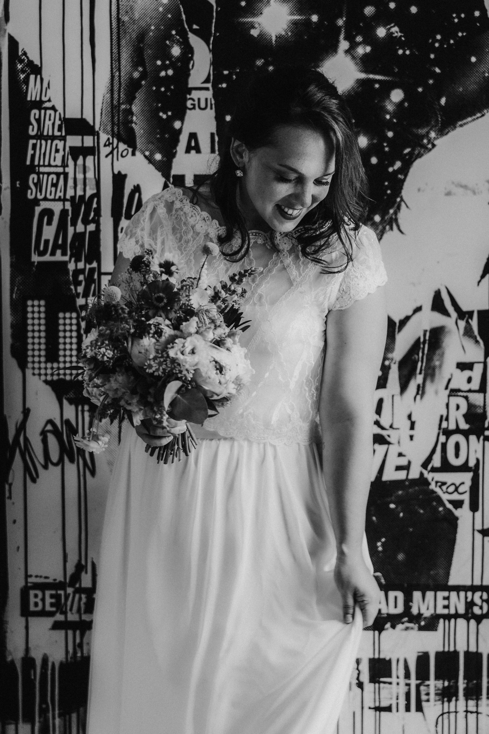 Daniel & Natalie’s Wedding at The Biscuit Factory 49 Bride in lace wedding dress holding a bouquet, smiling and looking down in front of a graphic wall.