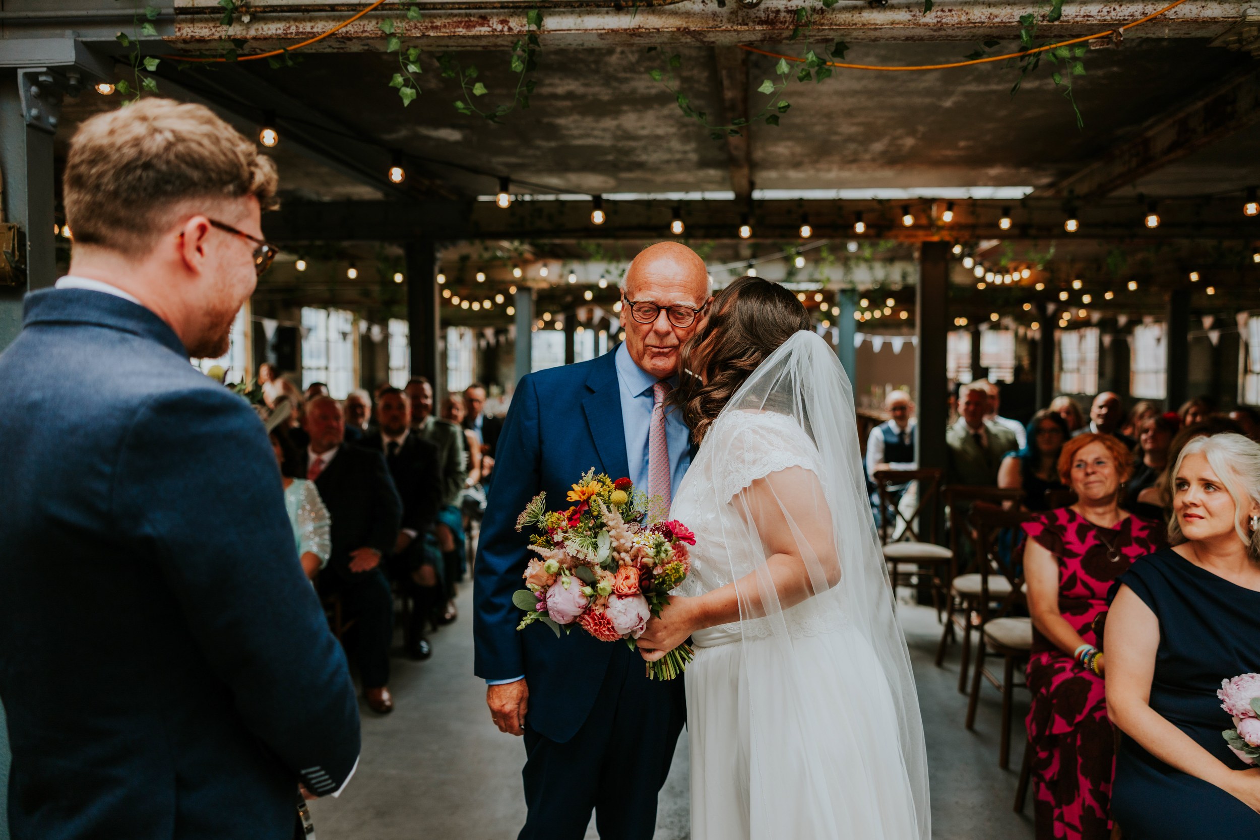 Daniel & Natalie’s Wedding at The Biscuit Factory 14 Bride in veil kisses an older man in a blue suit at the aisle, groom to left, guests and string lights in industrial venue.