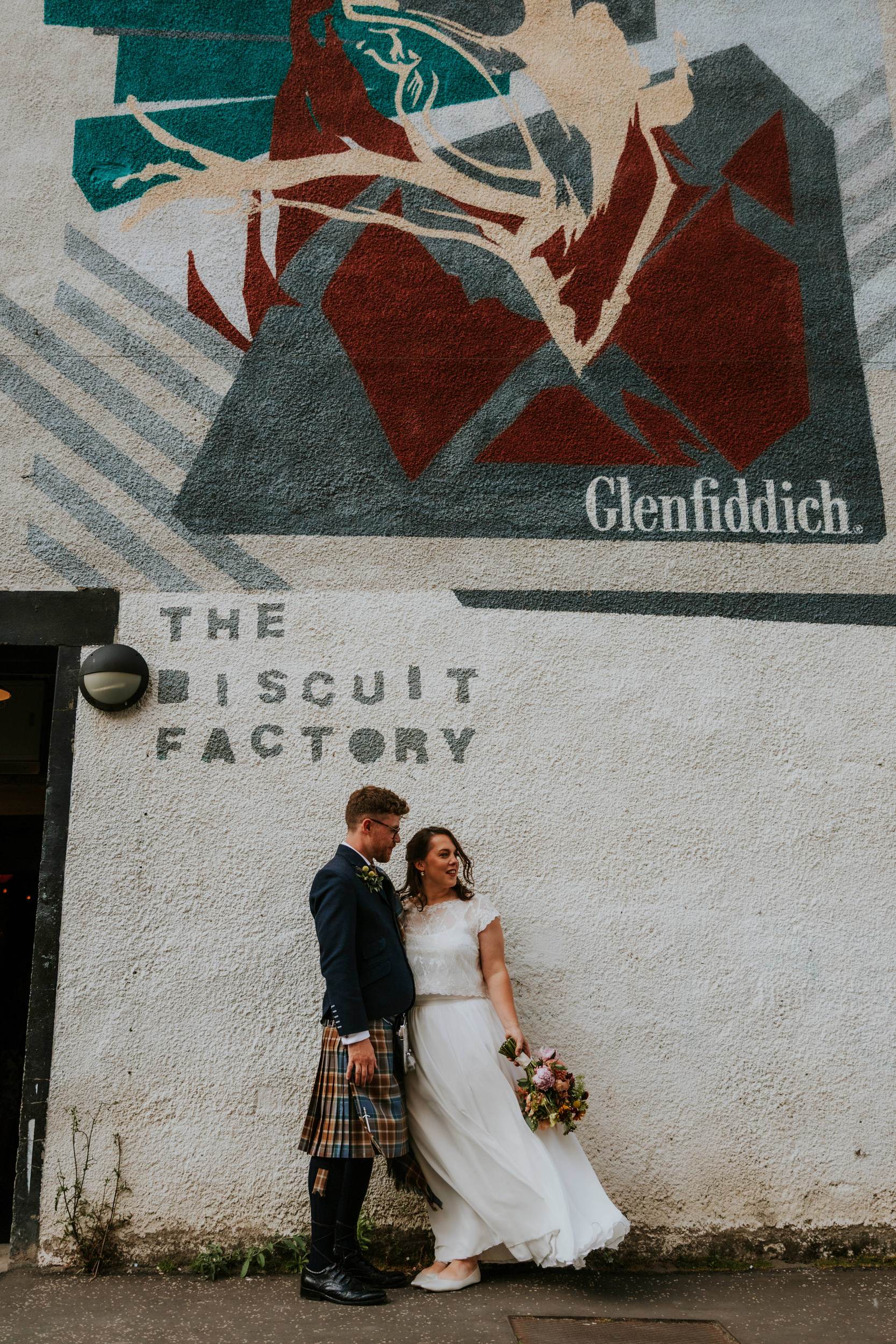 Daniel & Natalie’s Wedding at The Biscuit Factory 45 Bride in white dress and groom in kilt standing by The Biscuit Factory wall with mural, bride holding bouquet.