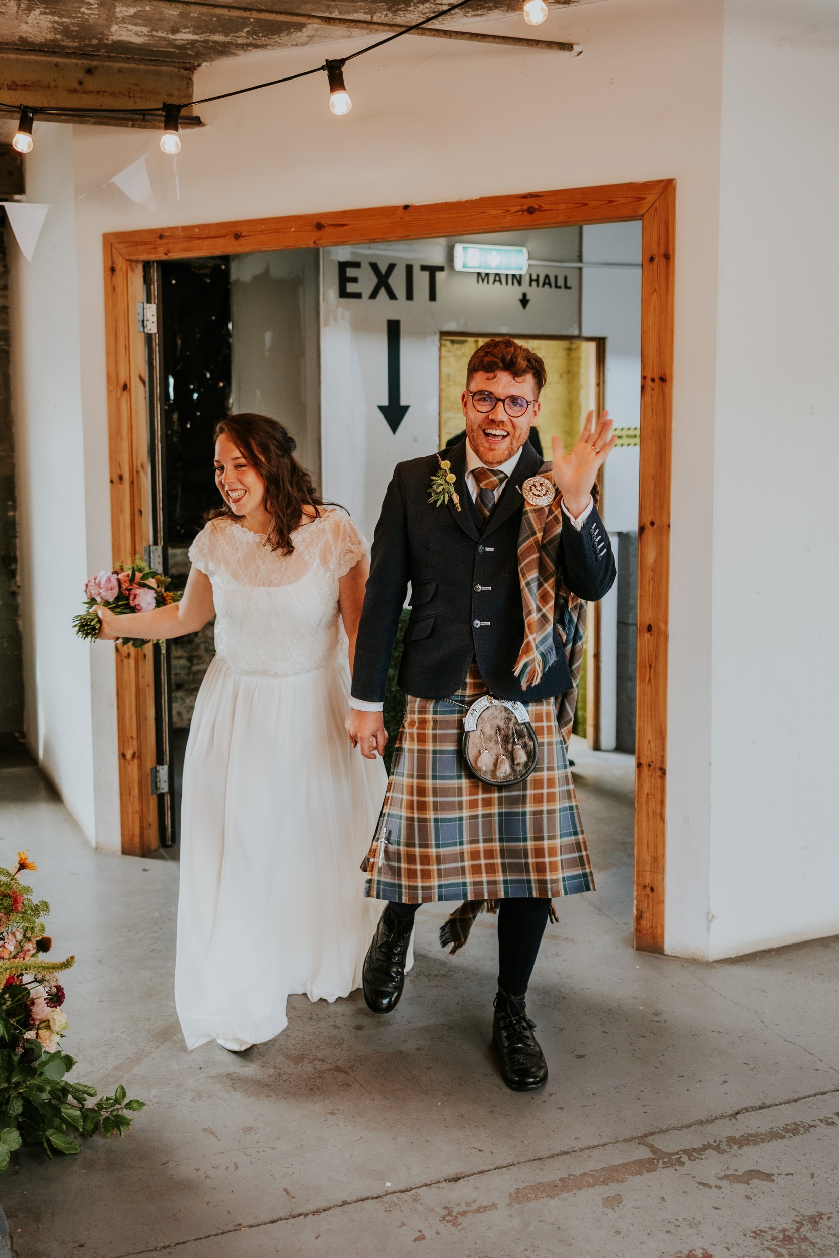 Daniel & Natalie’s Wedding at The Biscuit Factory 60 Bride in white dress and groom in tartan kilt holding hands as they enter through a doorway; groom waves.