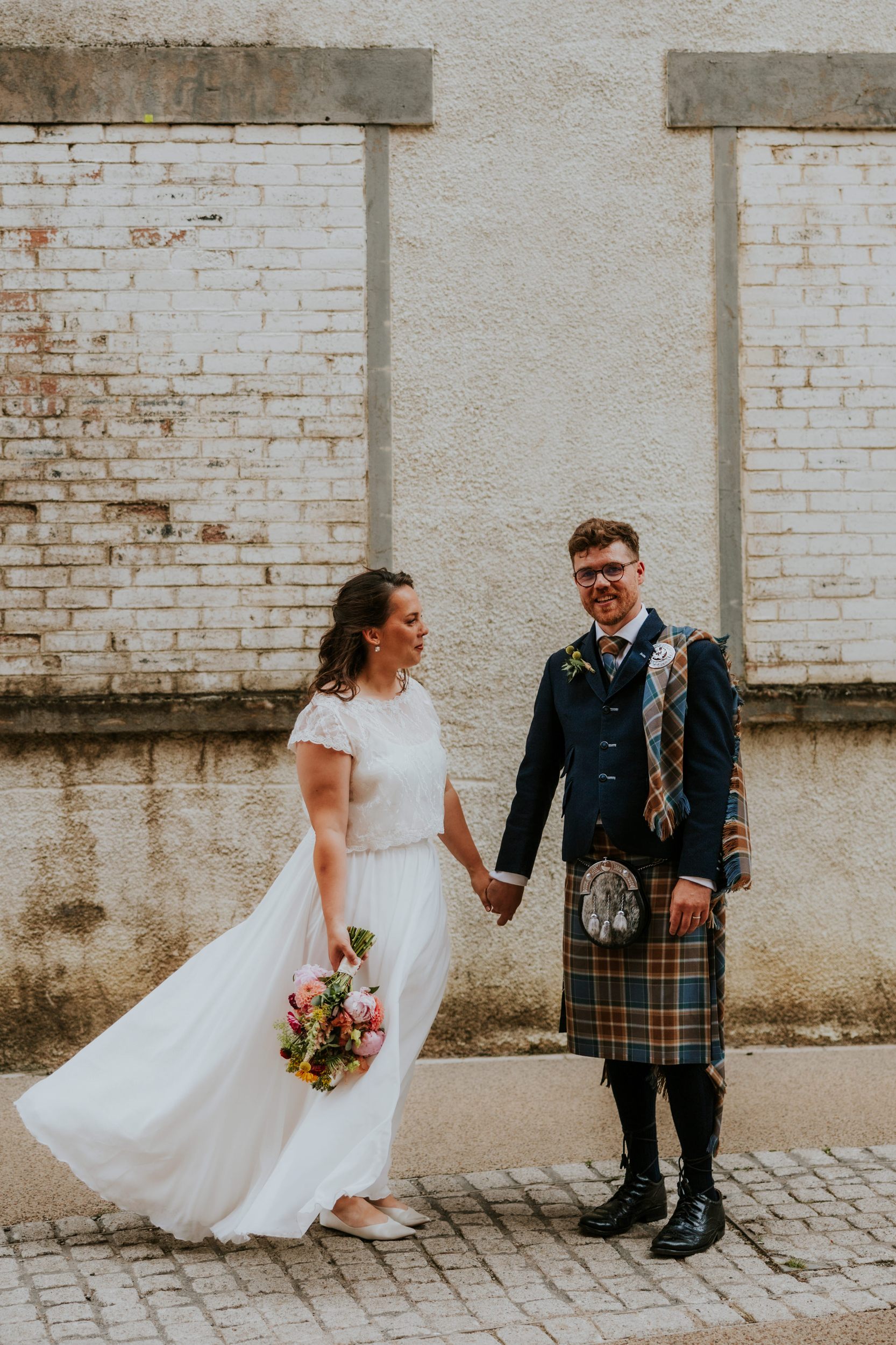 Daniel & Natalie’s Wedding at The Biscuit Factory 42 Bride in white dress with bouquet and groom in tartan kilt holding hands against a pale brick wall.