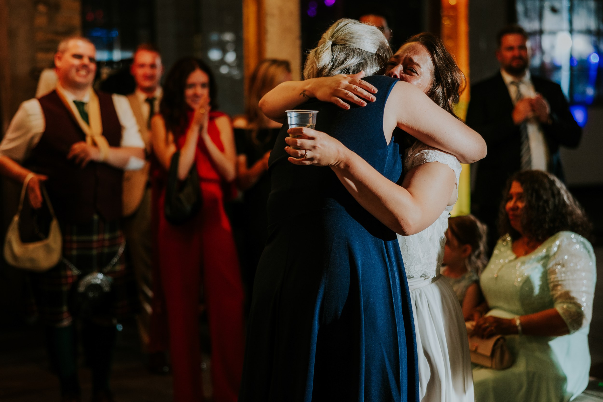 Daniel & Natalie’s Wedding at The Biscuit Factory 72 Bride in white hugs a guest in navy dress as guests clap at a wedding reception at The Biscuit Factory, Edinburgh.